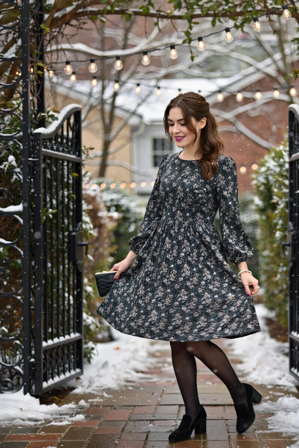 Woman twirling in a floral A-line midi dress flaring for movement, black tights & heels in a snowy garden under string lights