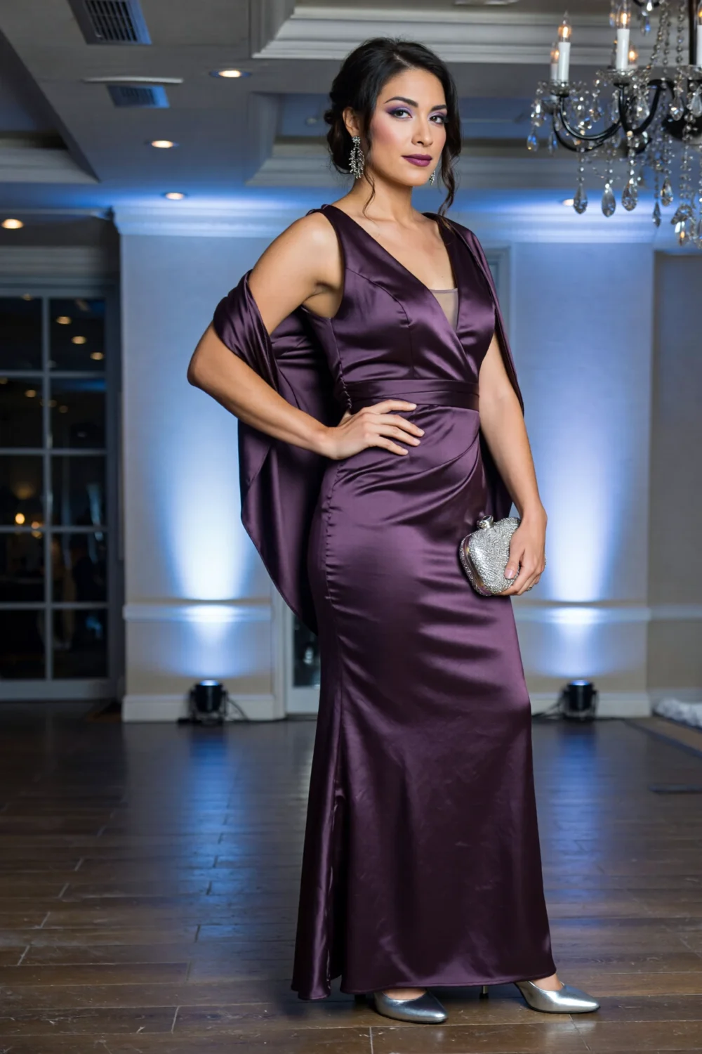Model in amethyst purple evening gown with draped shawl and glitter clutch beneath a chandelier, evoking ethereal winter elegance.