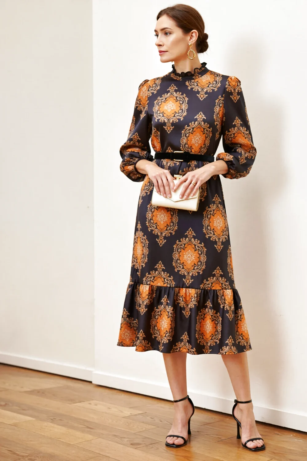 Model in charcoal Baroque midi with marigold florals, ruffled high neck, flounce hem, belted waist, black heels, ivory clutch