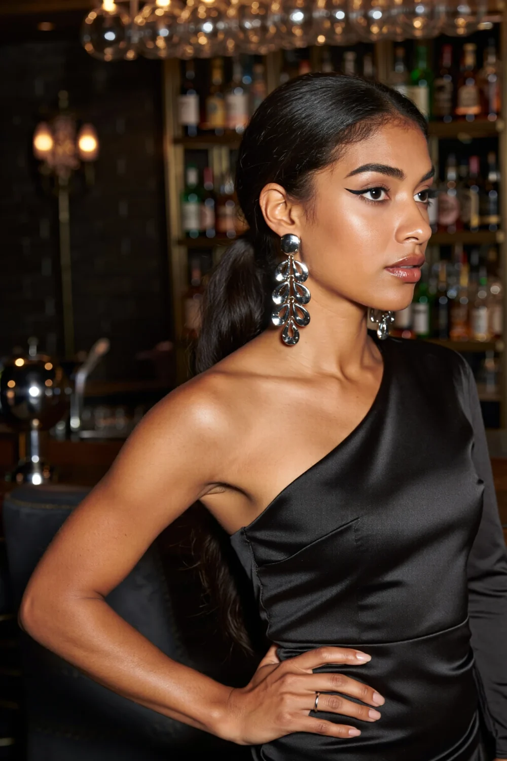 Model in one-shoulder black dress wearing sculptural silver chandelier earrings that float against a bare neckline, party-ready.