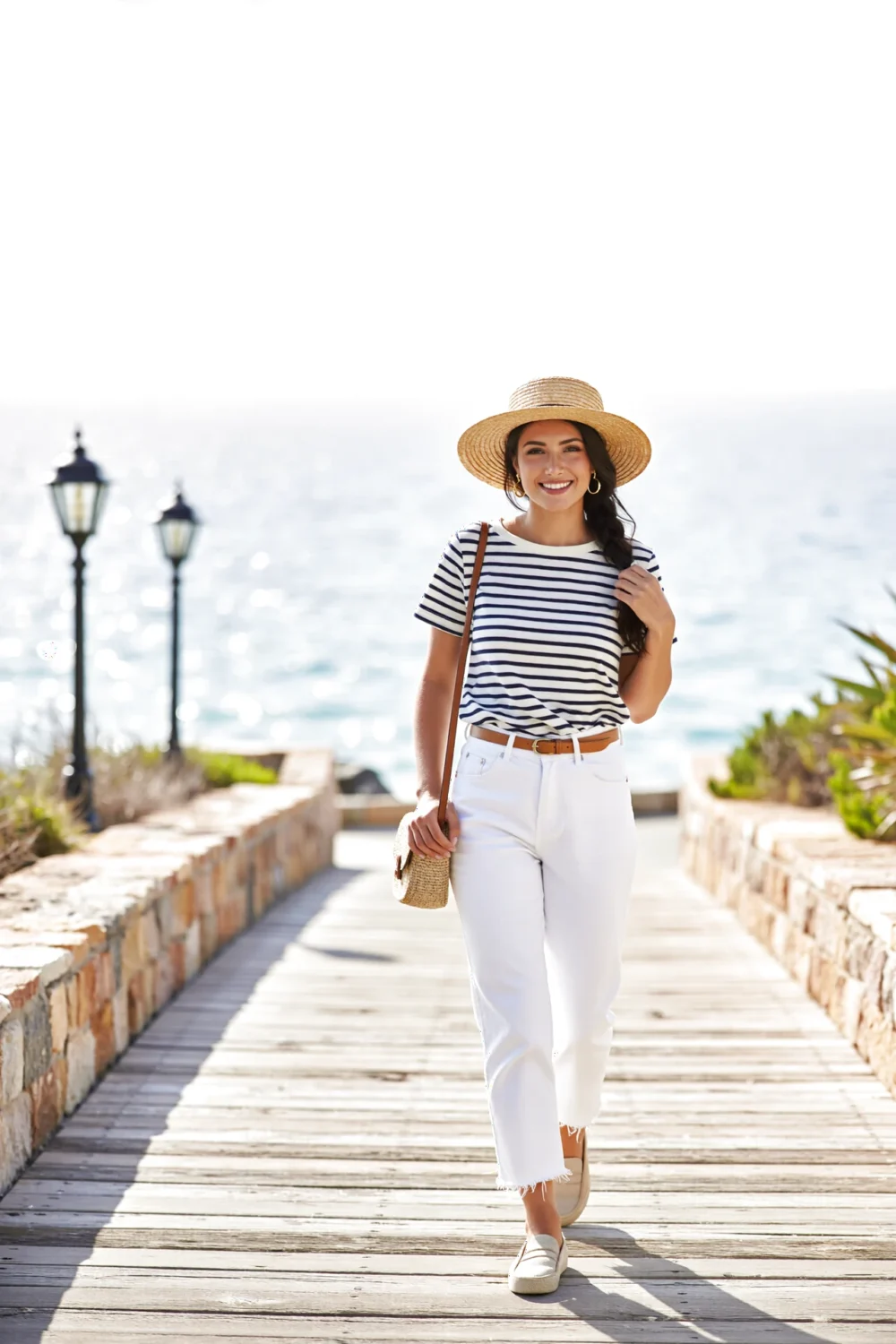 Smiling woman on boardwalk in a Breton navy-and-cream tee tucked into high white cropped jeans, straw boater and raffia crossbody