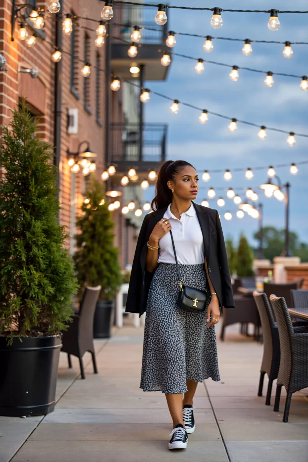 Woman wearing a polo tucked into a flowy midi skirt, sneakers and blazer, small crossbody and subtle jewelry under string lights