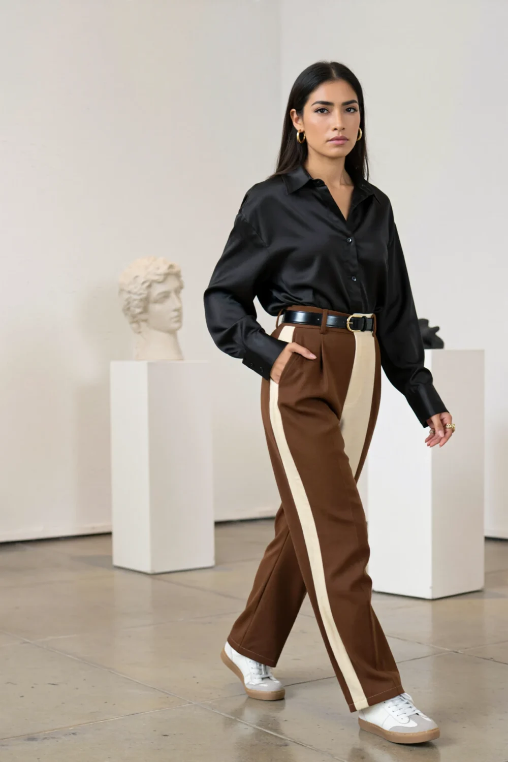 Model walks in gallery wearing oversized matte black shirt tucked into chocolate-and-ivory colorblock trousers, belt and sneakers