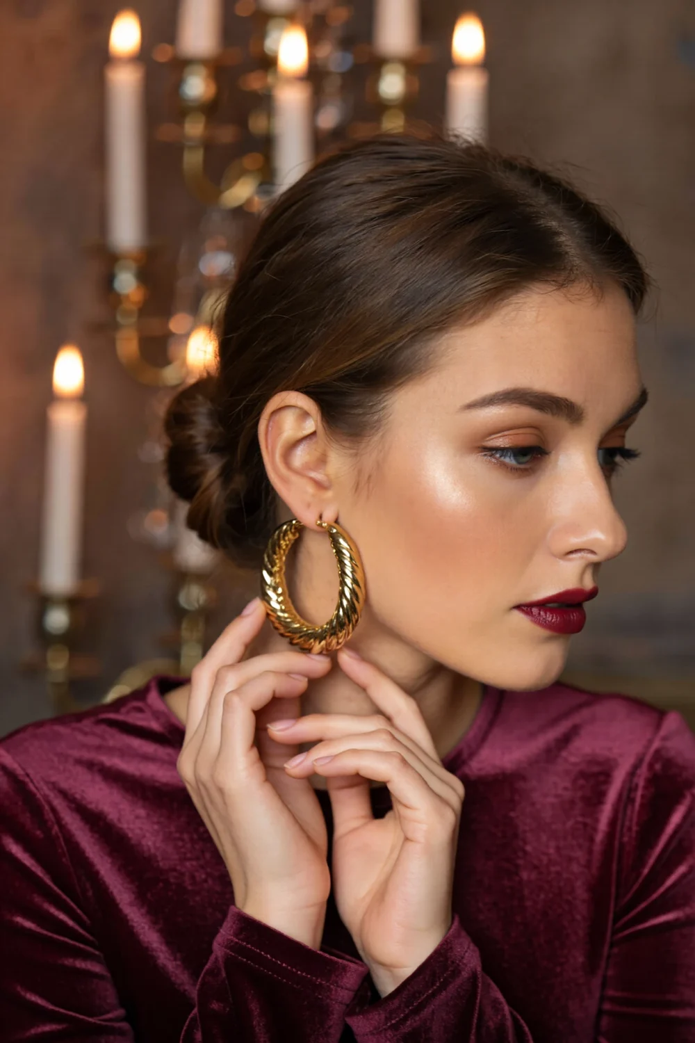 Woman in burgundy velvet top wearing a bold twisted gold hoop earring, a statement piece evoking layered mixed-metal styling.