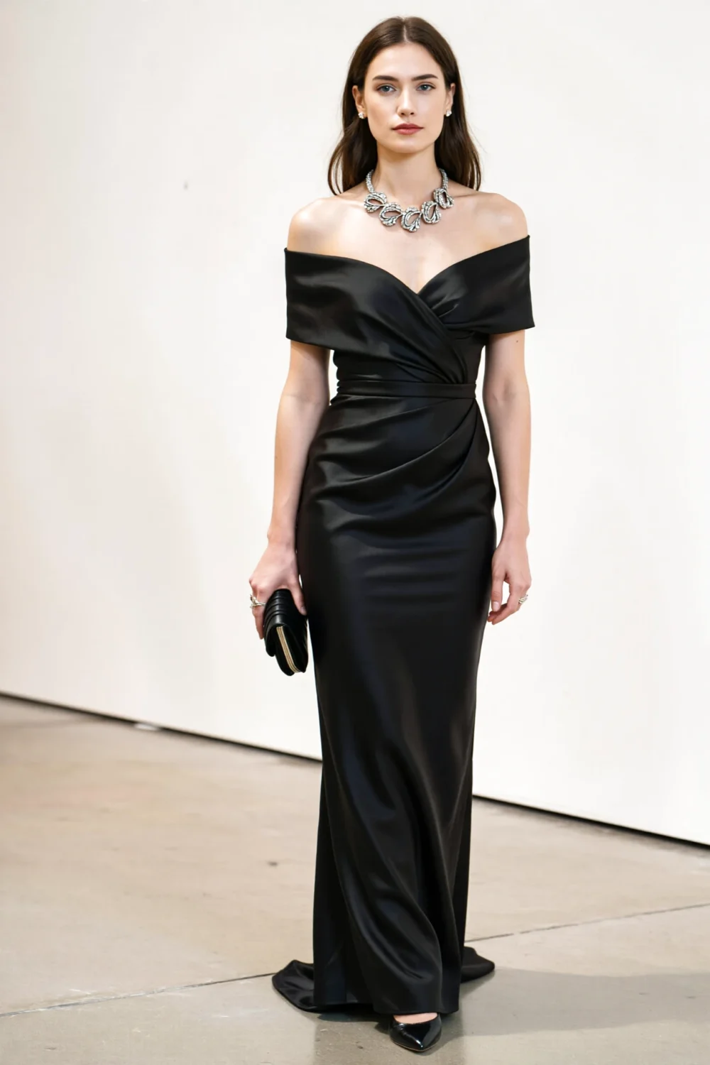 Model in a sleek black satin floor-length column gown with off-shoulder neckline, tailored waist, clutch and statement necklace.