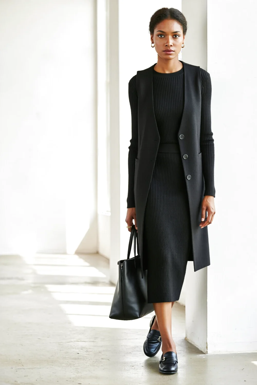 Model in black ribbed knit dress with long tailored vest, carrying a tote and wearing loafers for a minimal polished career look