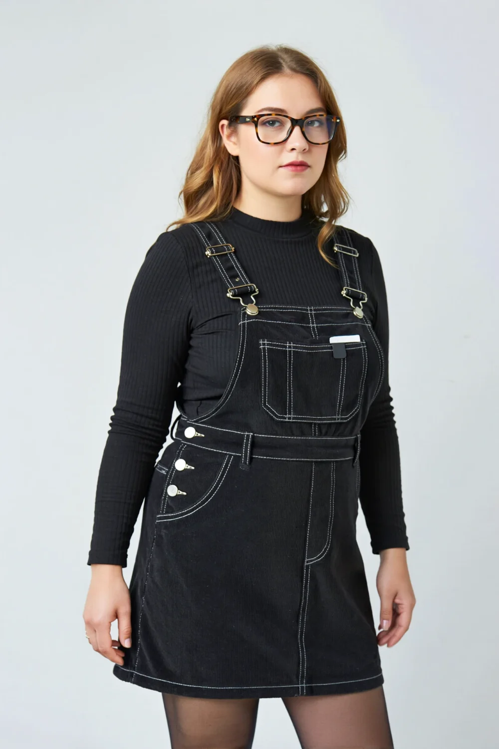 Black cord pinafore over a slim ribbed knit, styled with glasses for a playful schoolgirl-inspired seasonal outfit look