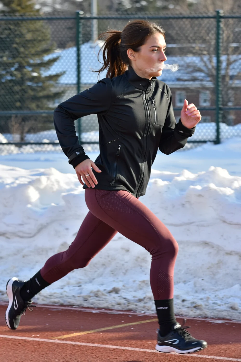 Runner in a cropped black windbreaker, maroon thermal leggings, low-profile gaiters and winter-traction shoes on a snowy track.