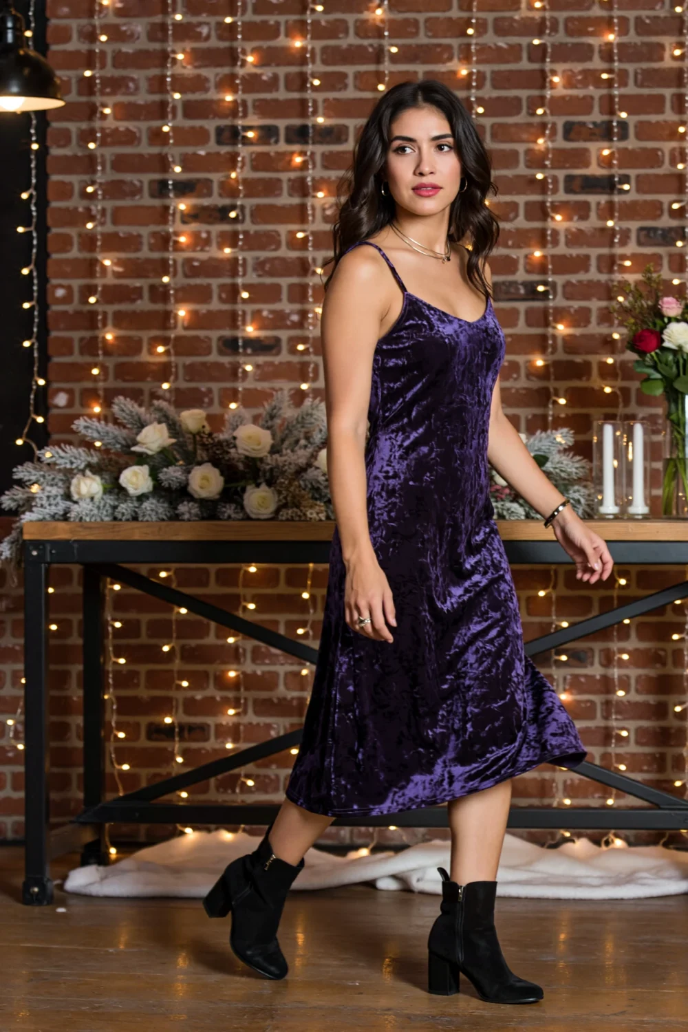 Woman in purple crushed velvet midi with thin straps and sheen, striding playfully in boots against a fairy-lit brick backdrop