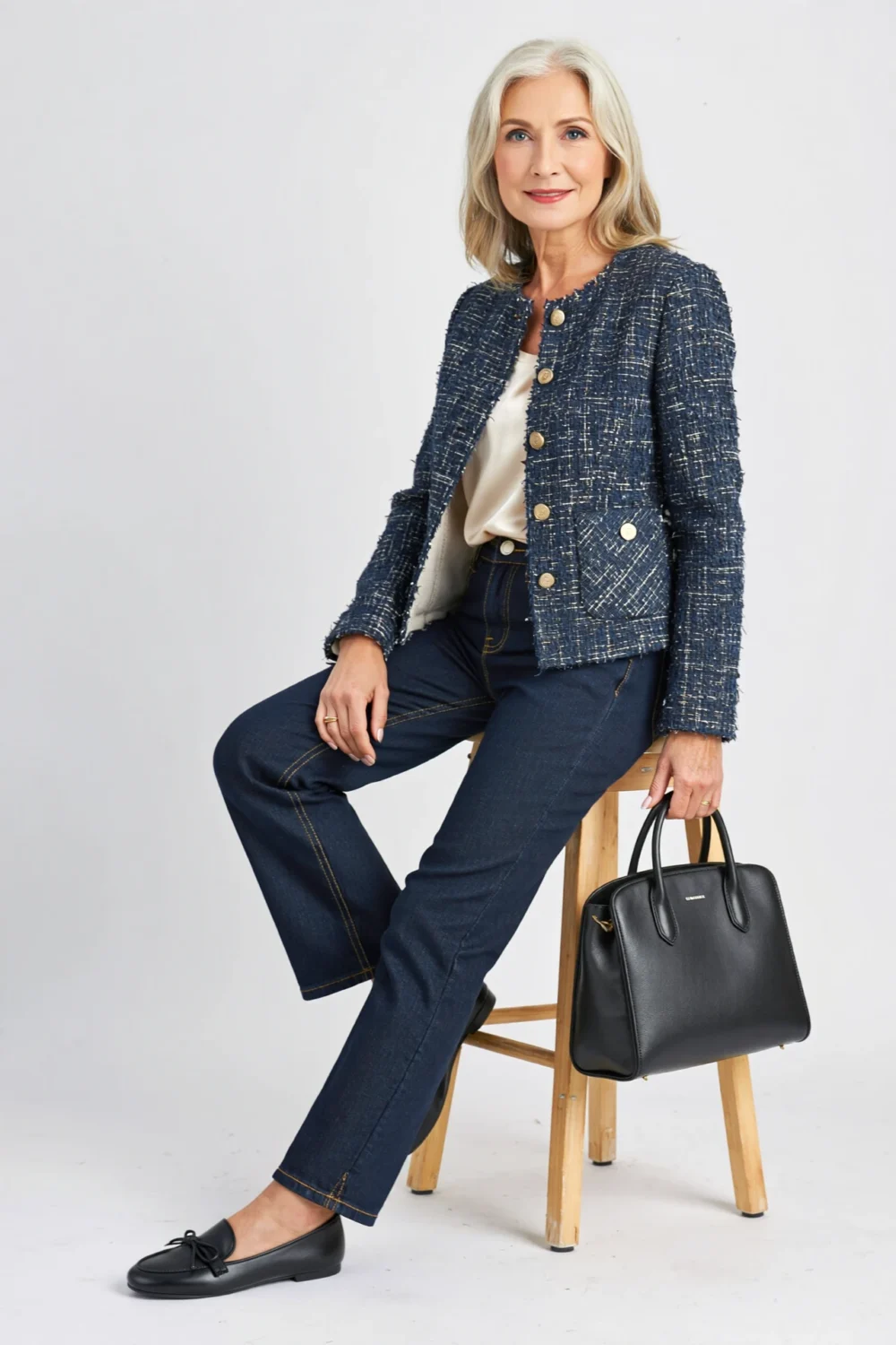Woman in dark pull-on straight jeans, silk tee and tweed jacket, with ballet flats and black top-handle bag, seated chicly