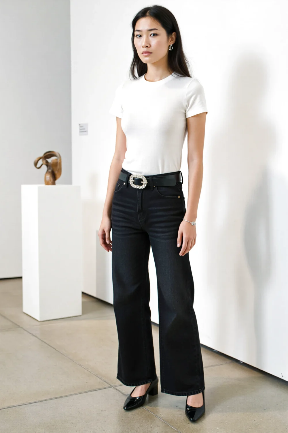 Model in a fitted white tee tucked into high-waisted wide-leg black jeans, snatched with a statement belt and pointed black heels.
