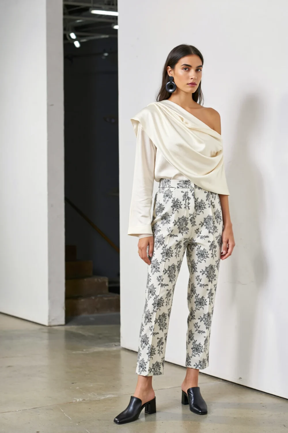 Gallery-ready outfit with sculptural ivory top, cropped floral trousers, and black heeled mules, styled for a modern opening look