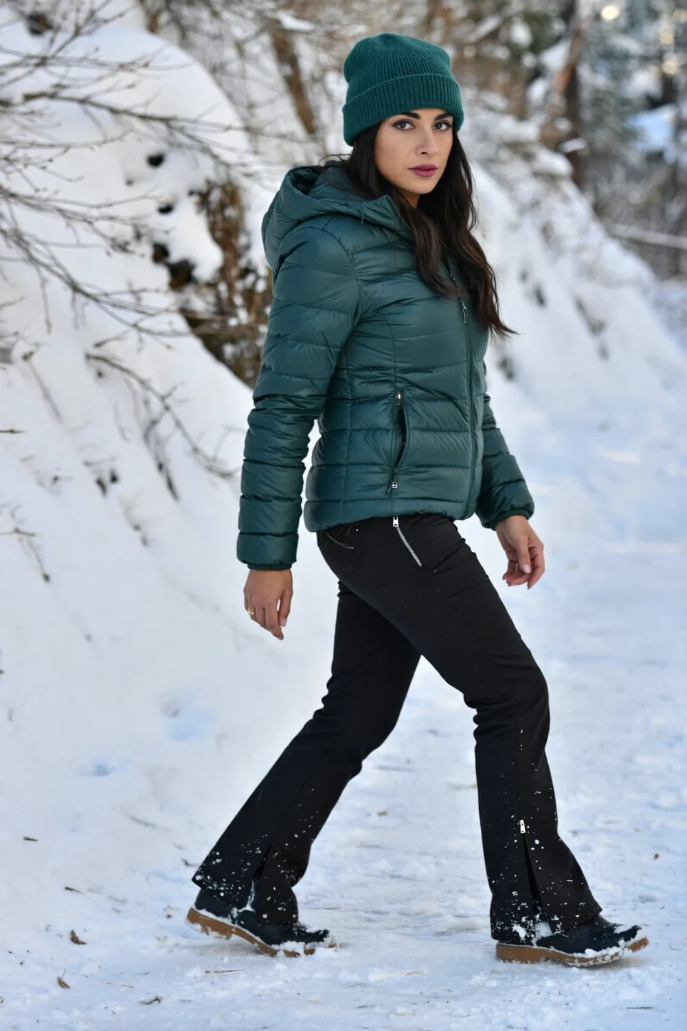 Woman in green puffer and beanie wearing high-rise black softshell pants with slight flare, zip pockets and winter boots in snow.