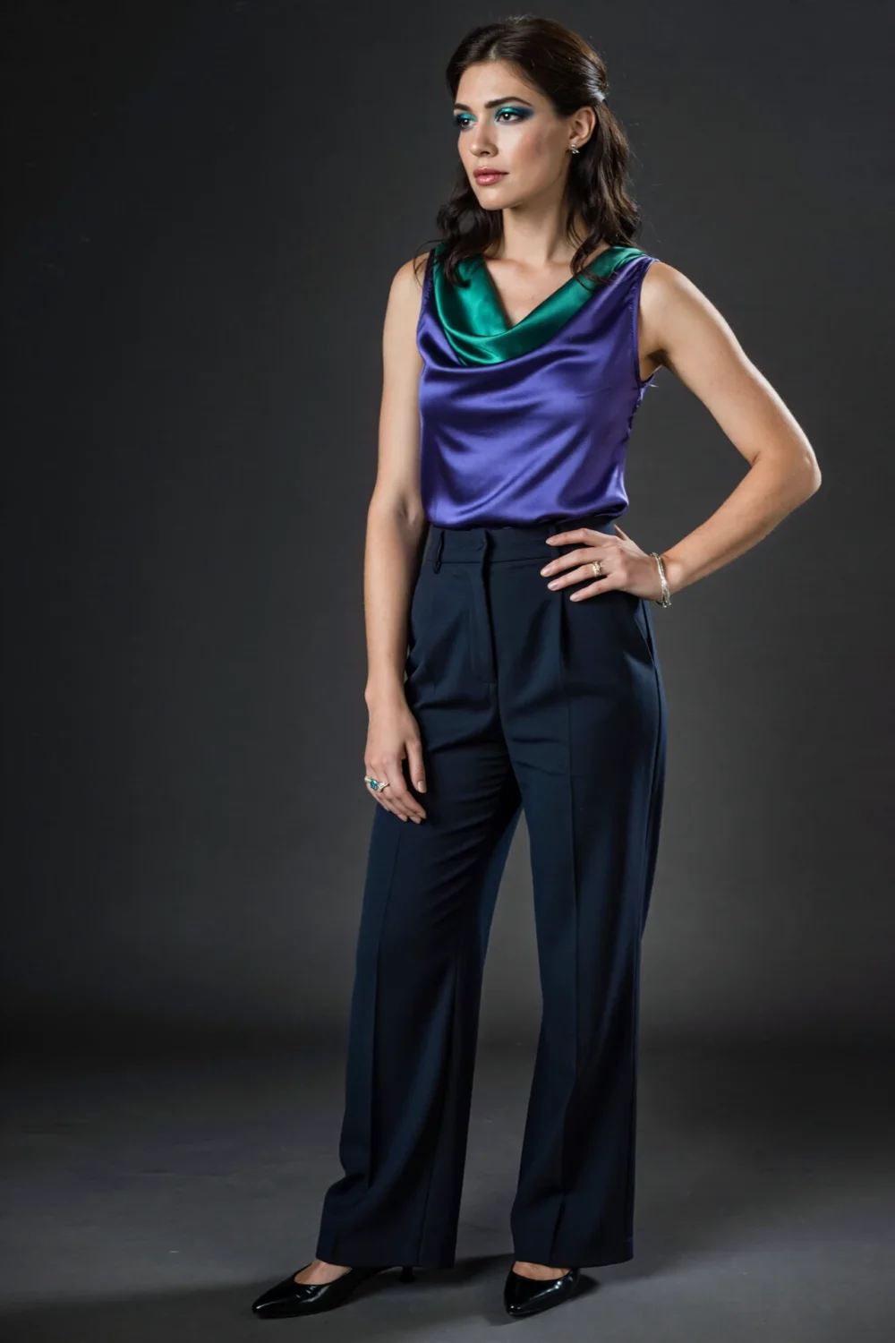 Woman wearing amethyst satin cowl-neck top with emerald drape and tailored navy trousers, minimal jewelry and sleek black heels