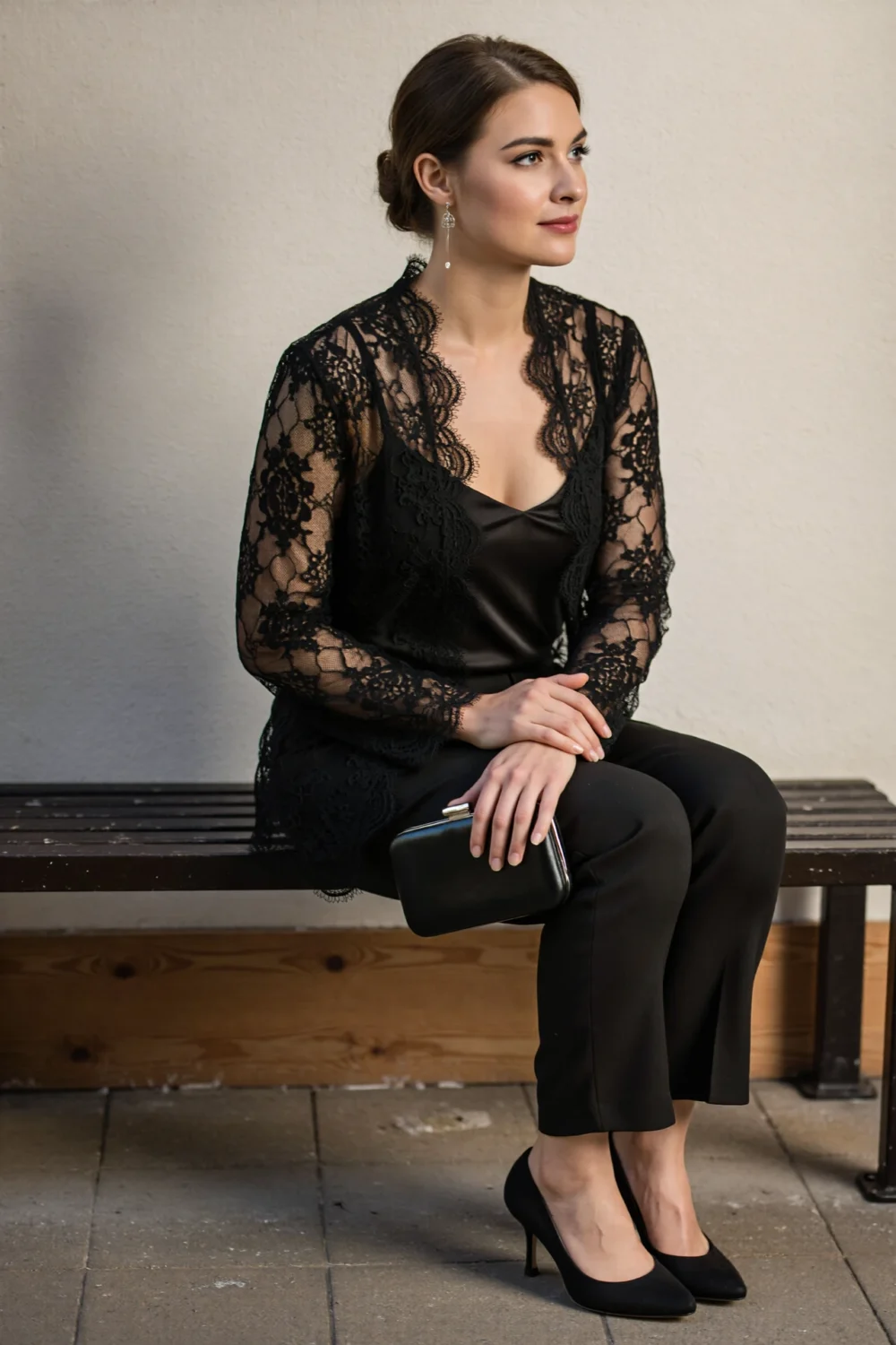 Woman seated in a black guipure lace blouse and tailored black trousers, delicate drop earrings, clutch and elegant black heels.