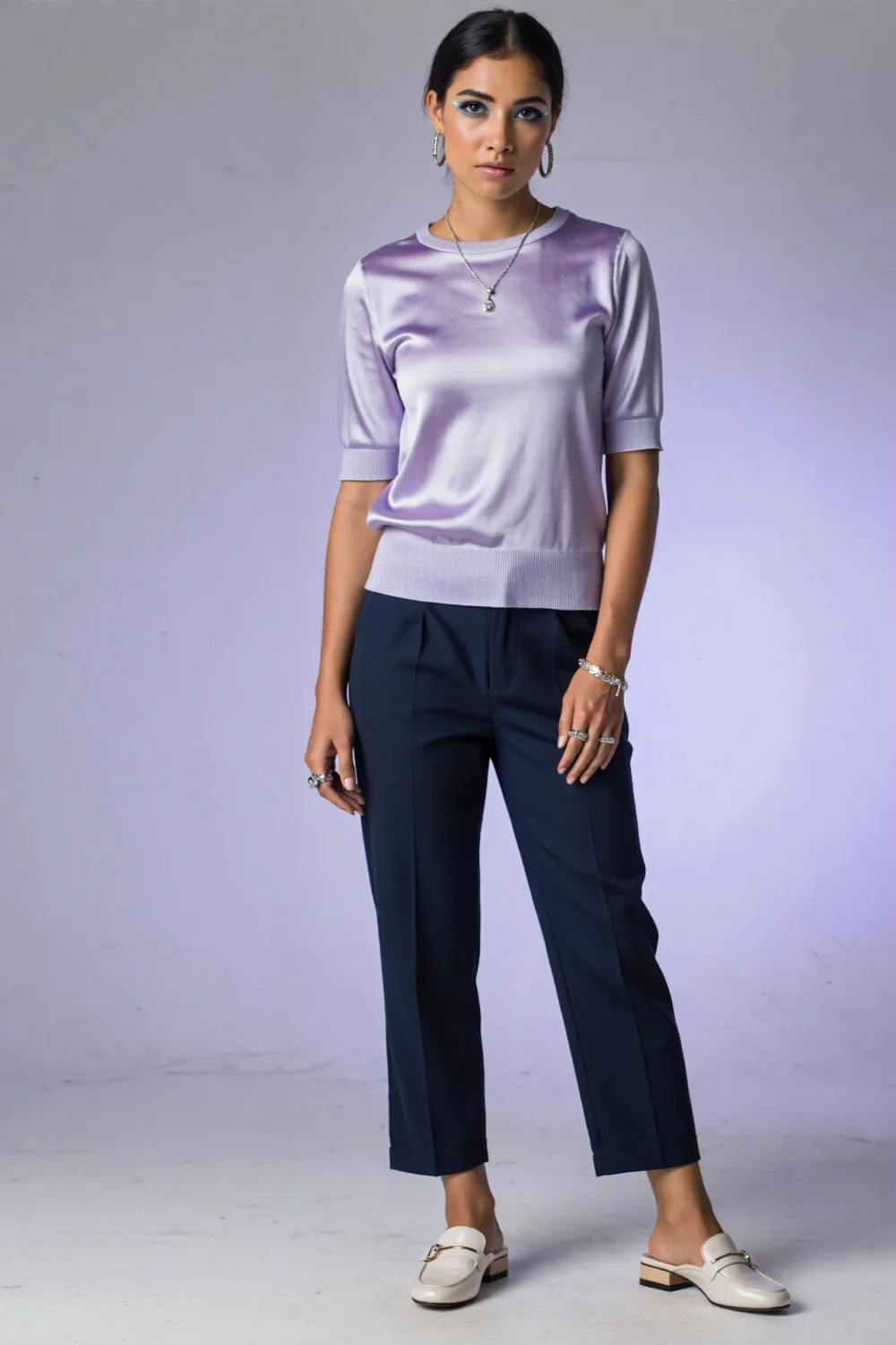 Model in a lilac satin top and navy tailored pants, accessorized with silver jewelry and white loafers against pale lilac backdrop