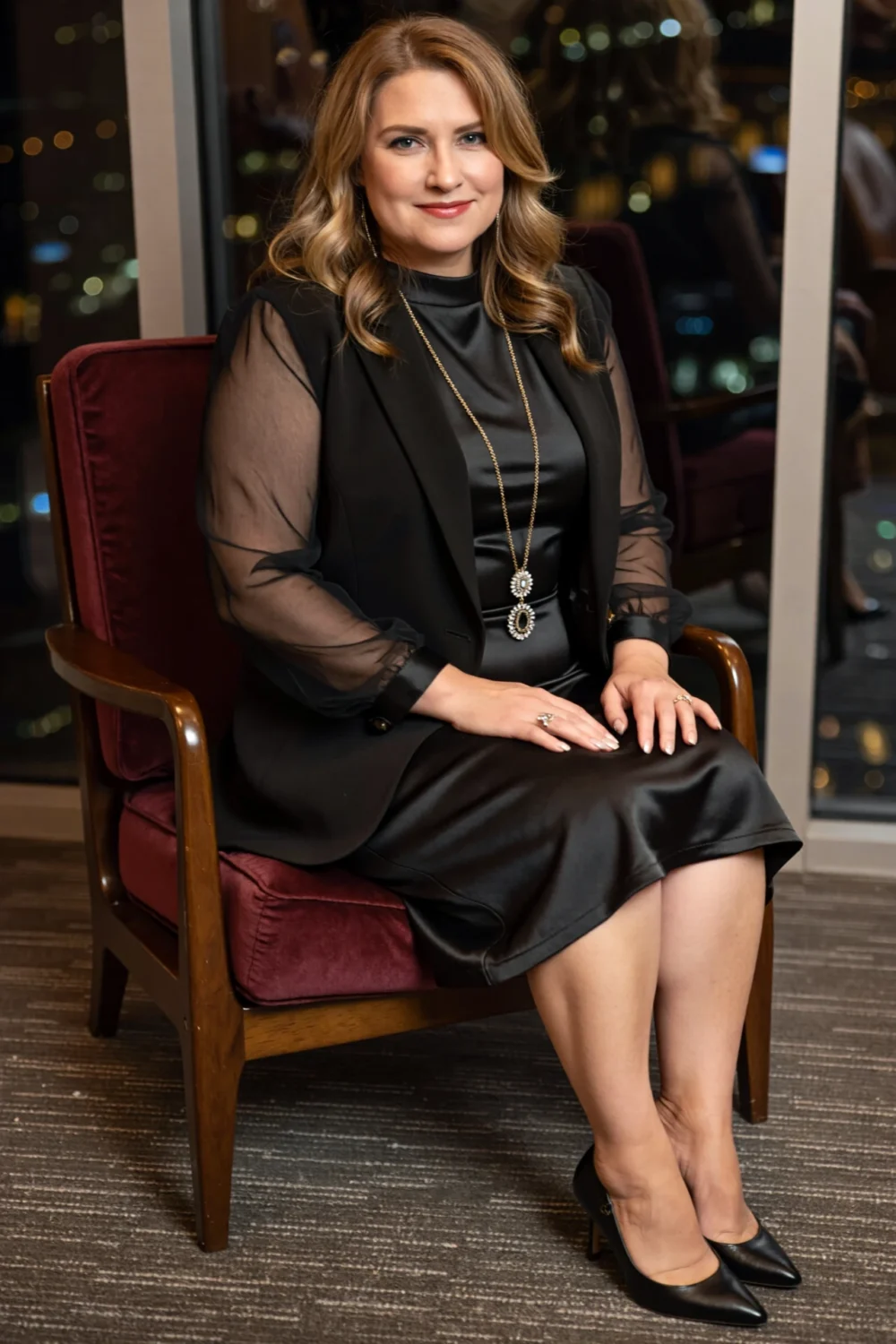 Seated woman in knee-length black A-line silk dress with sheer sleeves, layered blazer and long pendant necklace for evening.