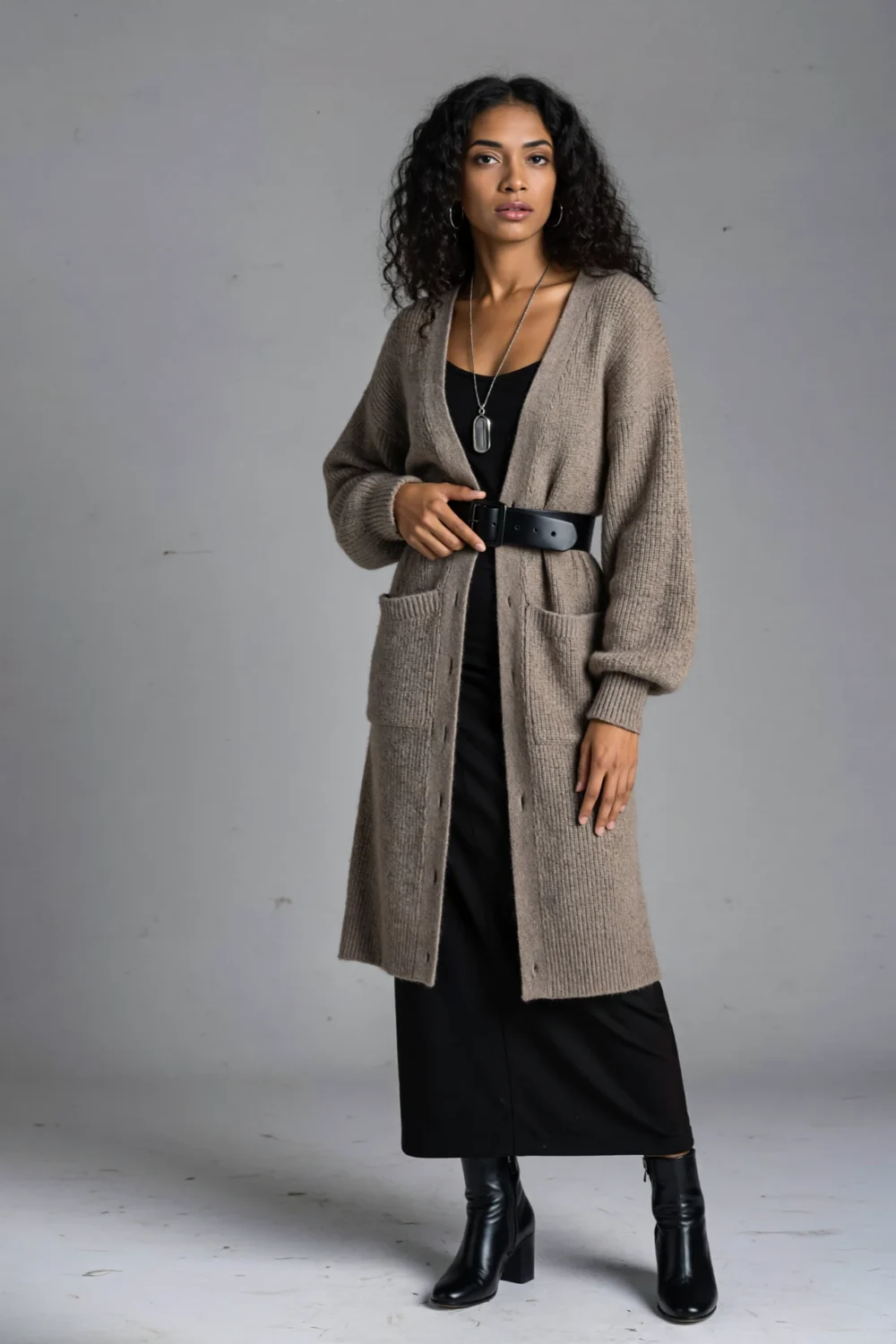 Model in a taupe longline belted cardigan over a black column skirt with heeled black boots and a long pendant, cozy tailored look