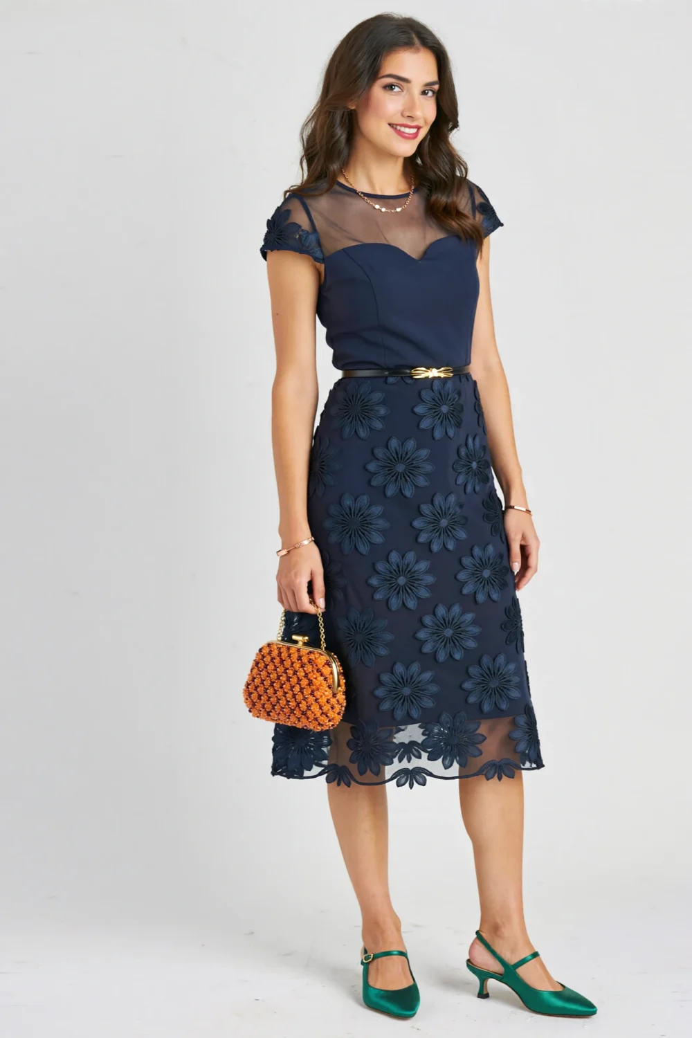 Model in navy 3D floral sheath with brushed-gold belt, emerald satin slingbacks, and marigold beaded minaudière for evening elegance
