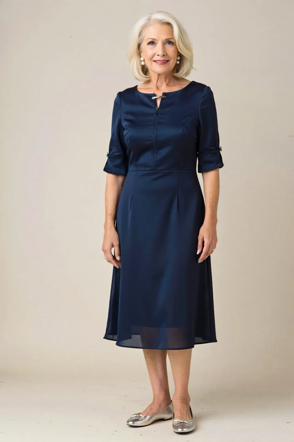 Elegant older woman in a navy tea-length crepe dress with soft sleeves, pearl studs, and metallic flats, standing poised indoors