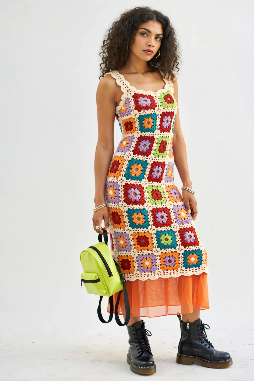 Model in colorful granny-square crochet midi dress with tangerine slip, neon mini backpack, and black combat boots on white background