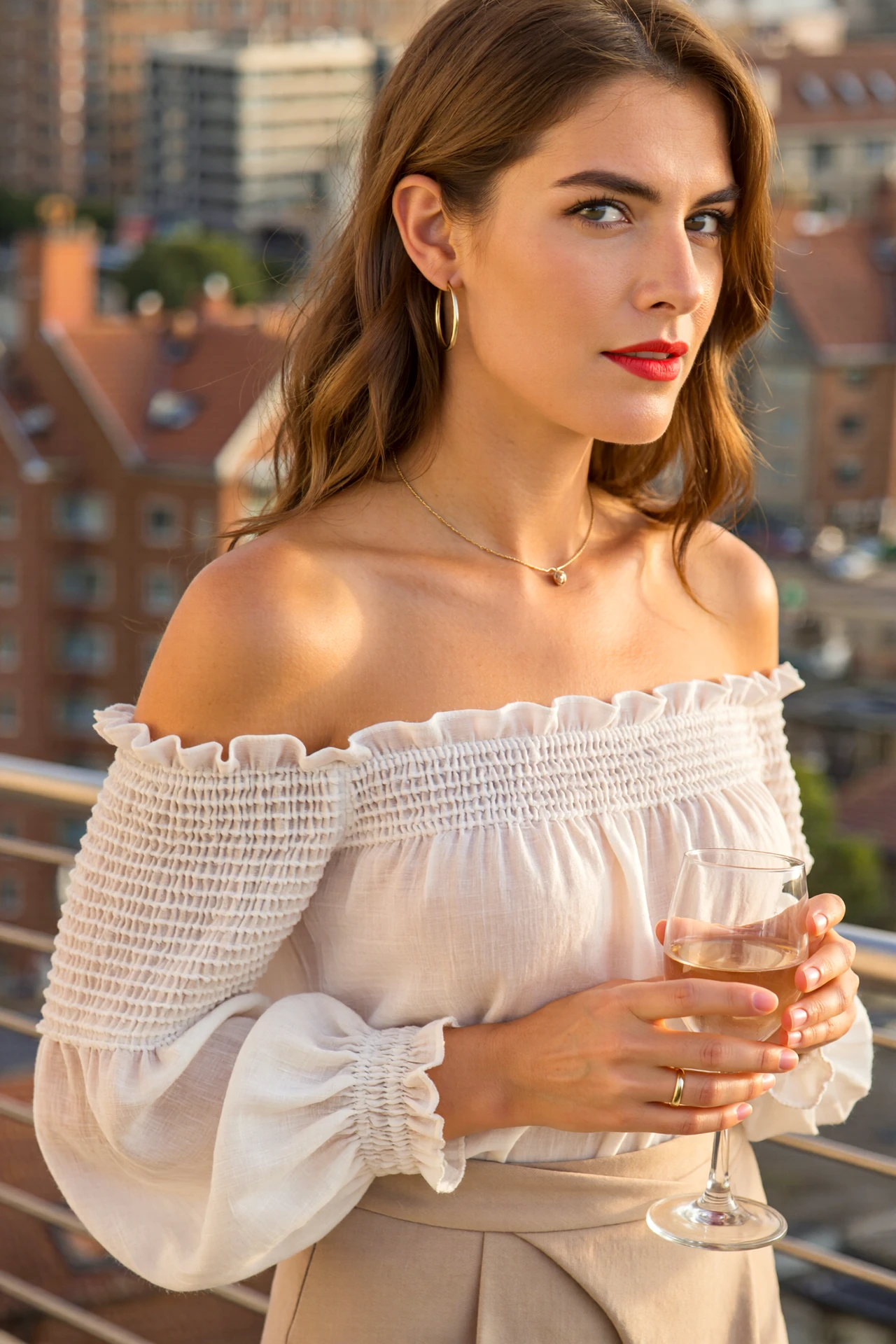 Woman in an off-shoulder cotton-voile blouse with pintucks and smocked cuffs, sipping aperitivo on a sunset terrace.