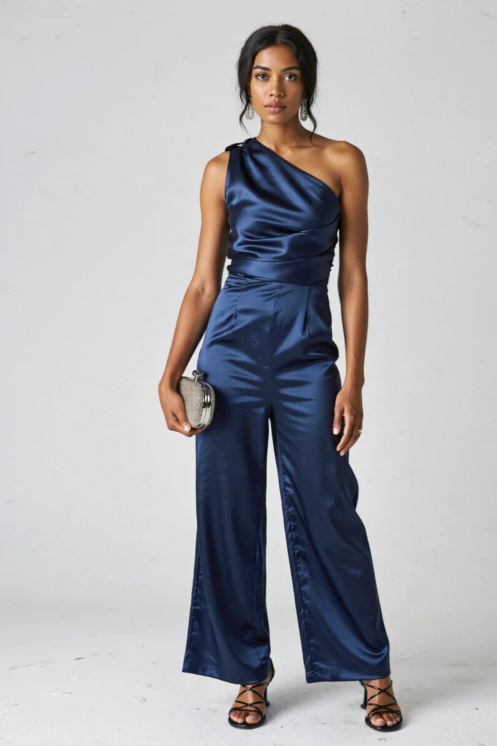 Navy one-shoulder satin jumpsuit with sculptural drape and tailored waist, styled with strappy sandals and metallic clutch—elegant