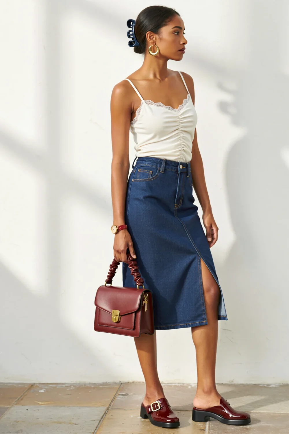 Woman in ruched white camisole and indigo denim midi skirt with burgundy mules, doctor-style bag, and gold accents, vintage chic