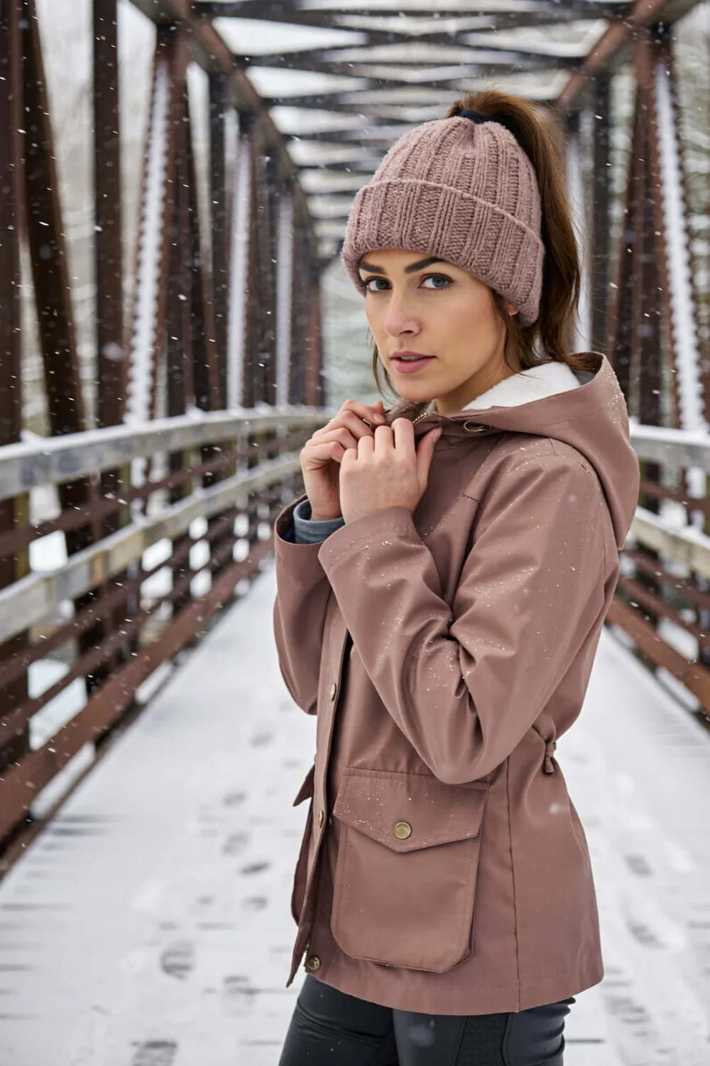 Woman on snowy bridge wearing mauve ponytail beanie with high ponytail, hood-friendly, layers over merino base under hooded shell