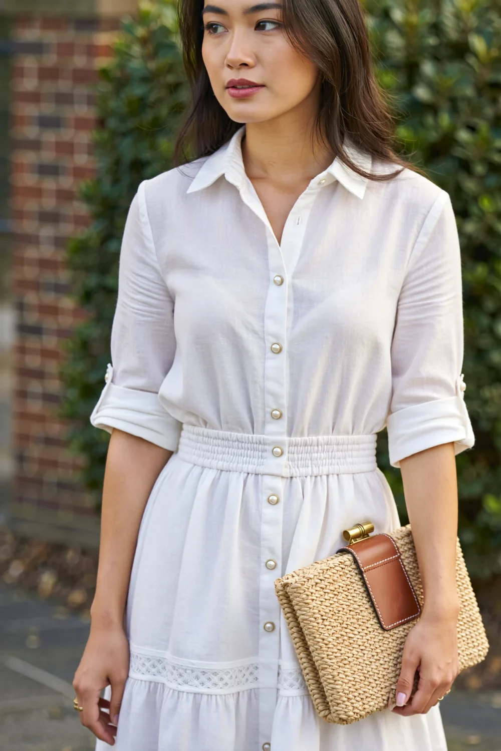 Woman in a crisp white poplin shirt dress with belted waist, holding a woven raffia clutch for a relaxed daytime look
