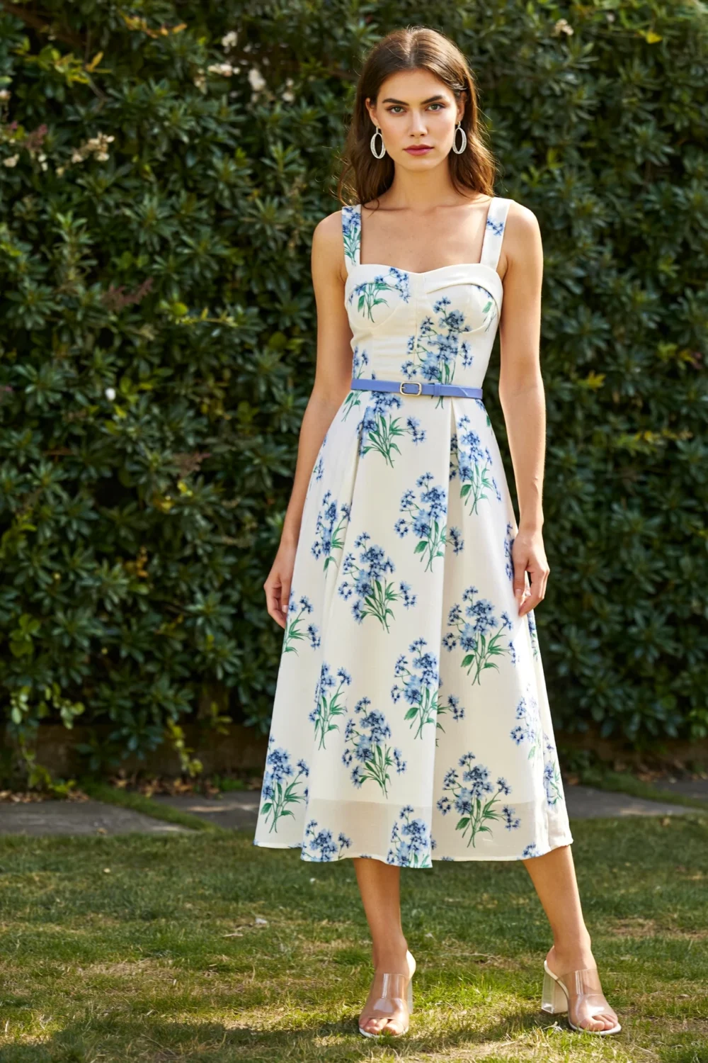 Model in ivory cotton-faille midi dress with blue floral print, square neckline, wide straps, and periwinkle belt against greenery.