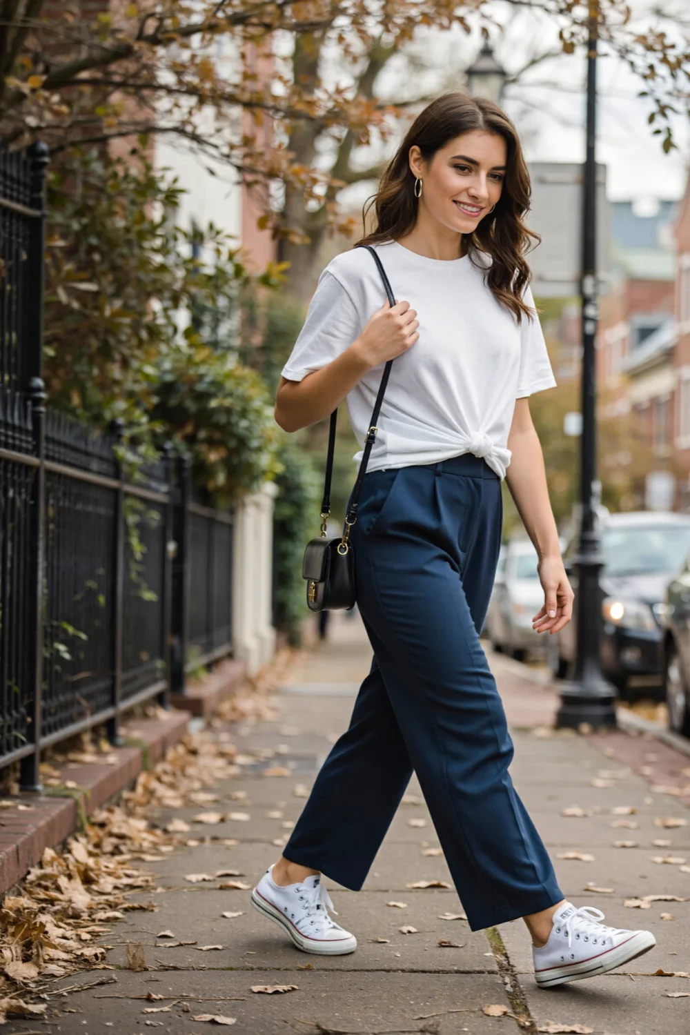 Woman in a knotted white tee, navy wide-leg pants and sneakers walks city sidewalk with crossbody bag and simple hoop earrings.