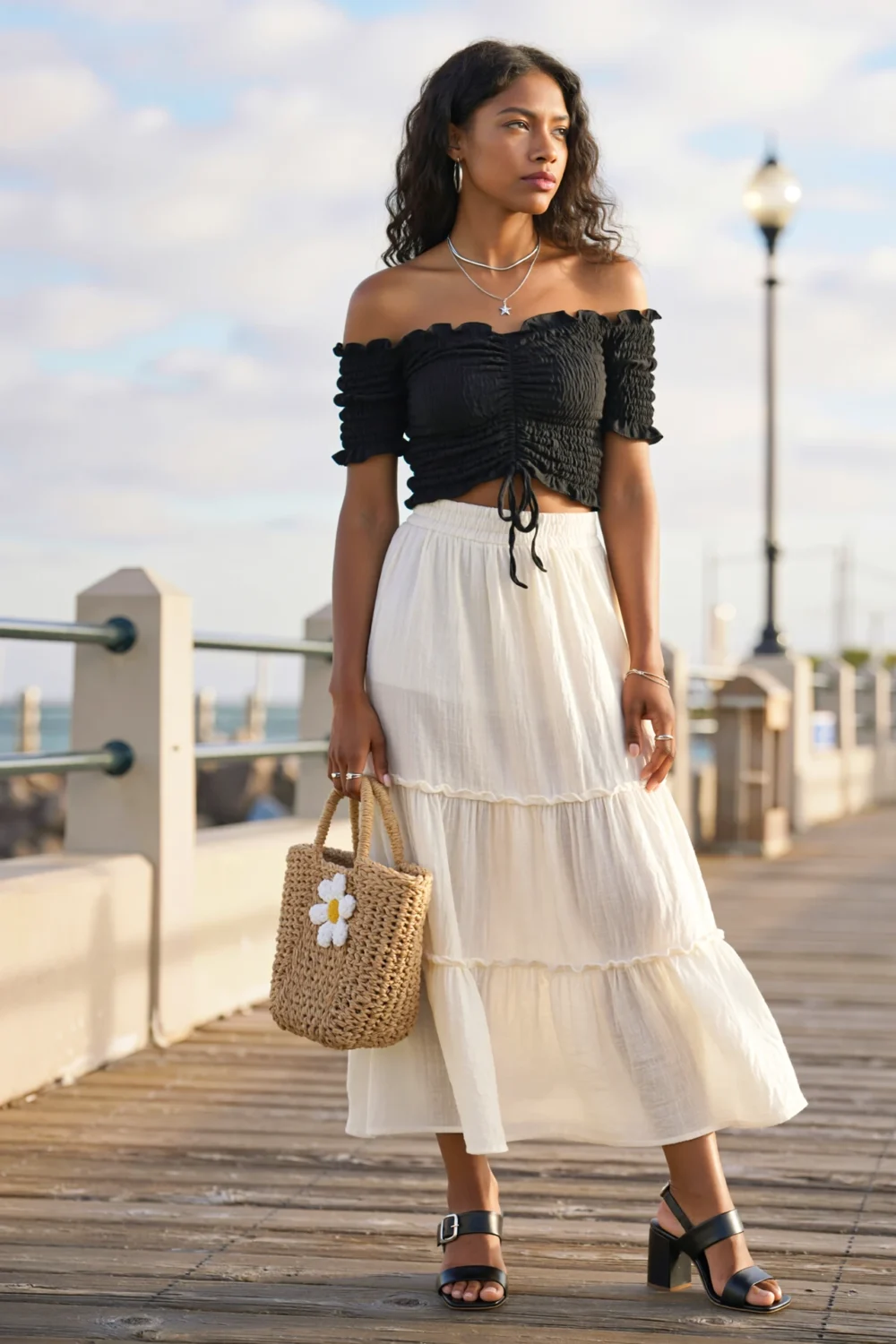 Woman in ruched black off-shoulder top with tiered white maxi skirt, black heels and raffia tote on a boardwalk, editorial chic