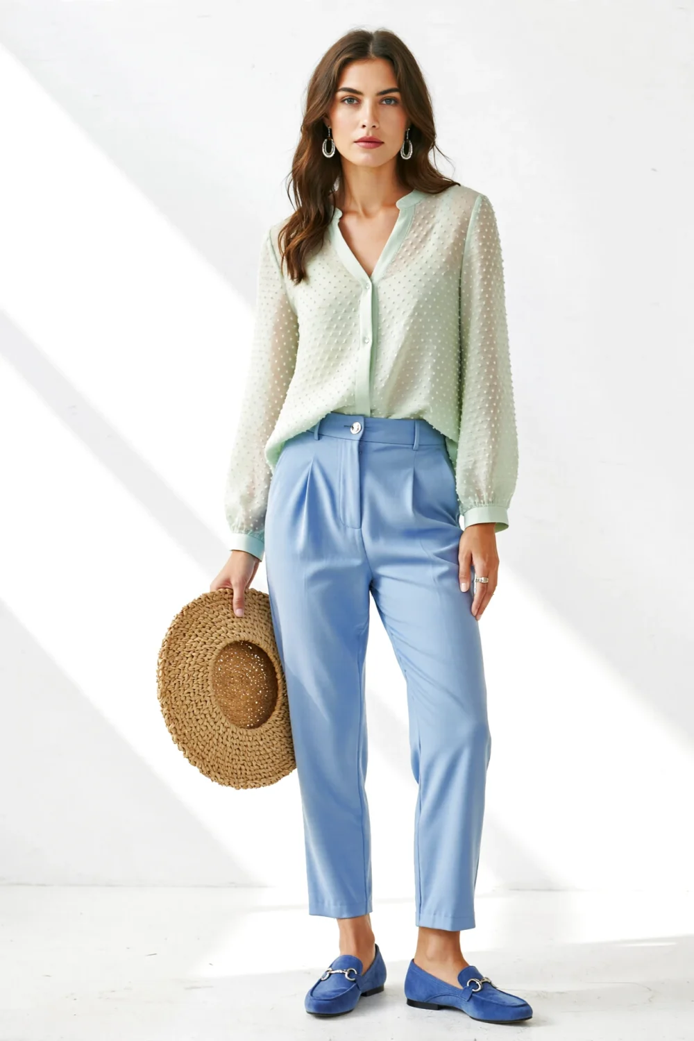 Model in sage blouse with sky blue tapered pants, silver earrings, straw hat, and blue loafers for airy summer styling