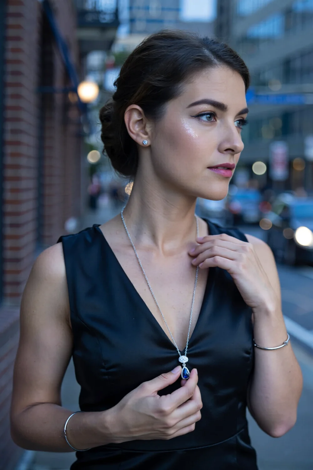 Woman in black dress wearing a mid-length silver chain with sapphire pendant, silver studs and slender bracelet, serene elegance.