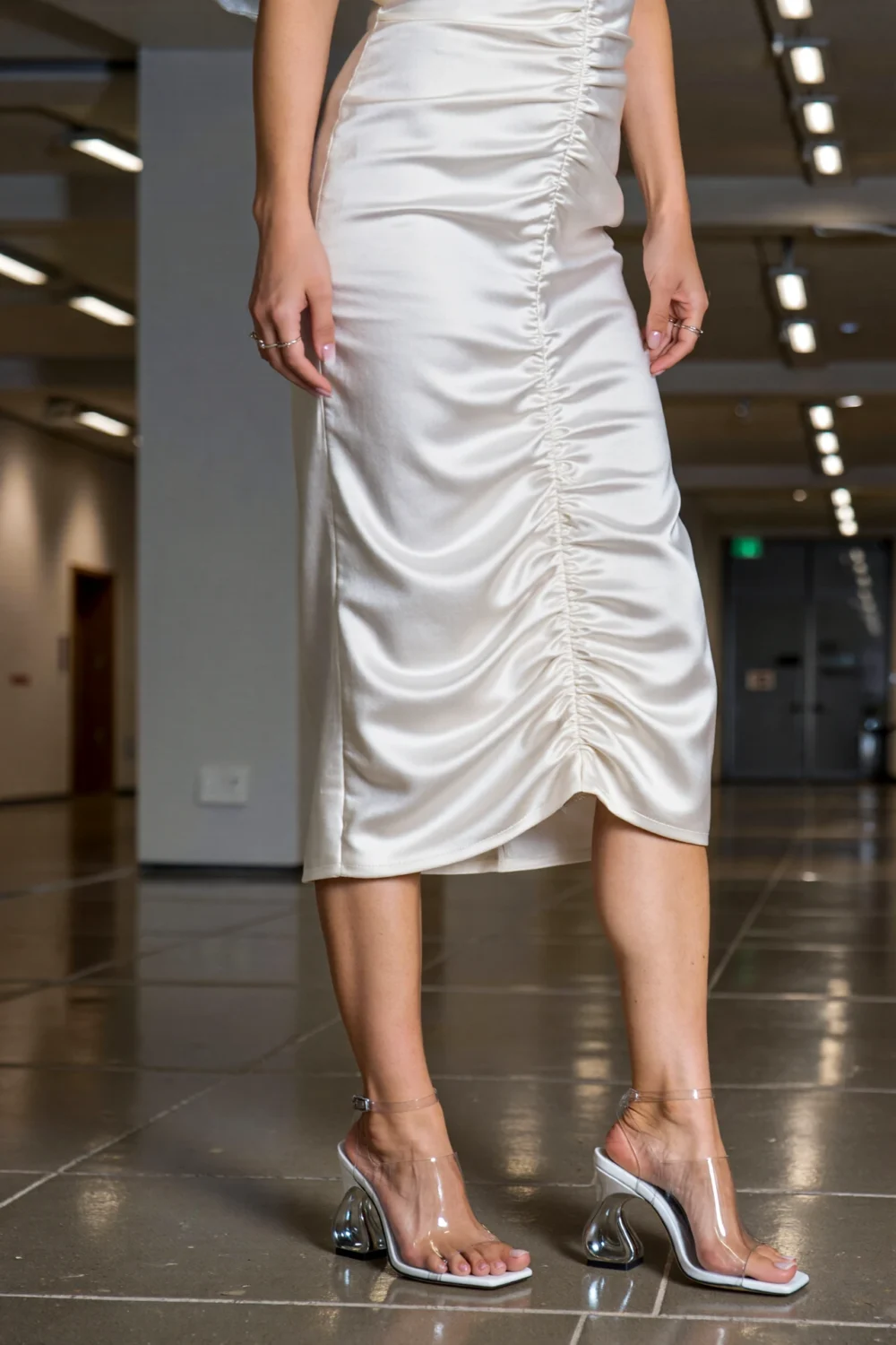 White satin midi dress with sculptural metallic heels and clear straps, creating a glossy monochrome couture look on the runway floor