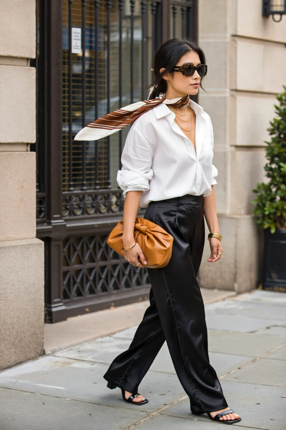 Woman in a crisp white shirt, black satin trousers, silk scarf, and square-toe sandals, carrying a tan clutch on city street