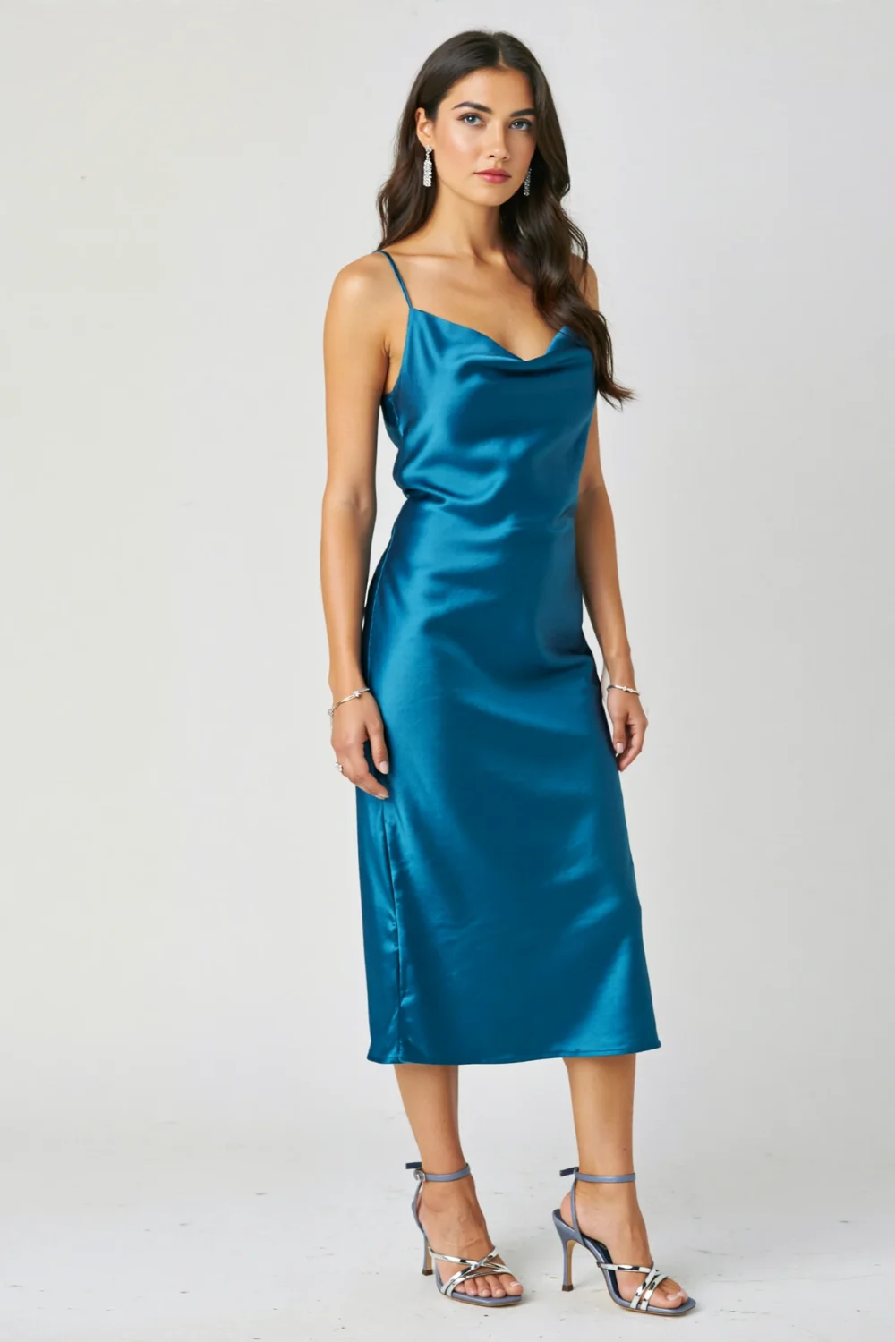Model in a teal satin midi dress with silver strappy heels and crystal earrings, creating a sleek, modern evening look.