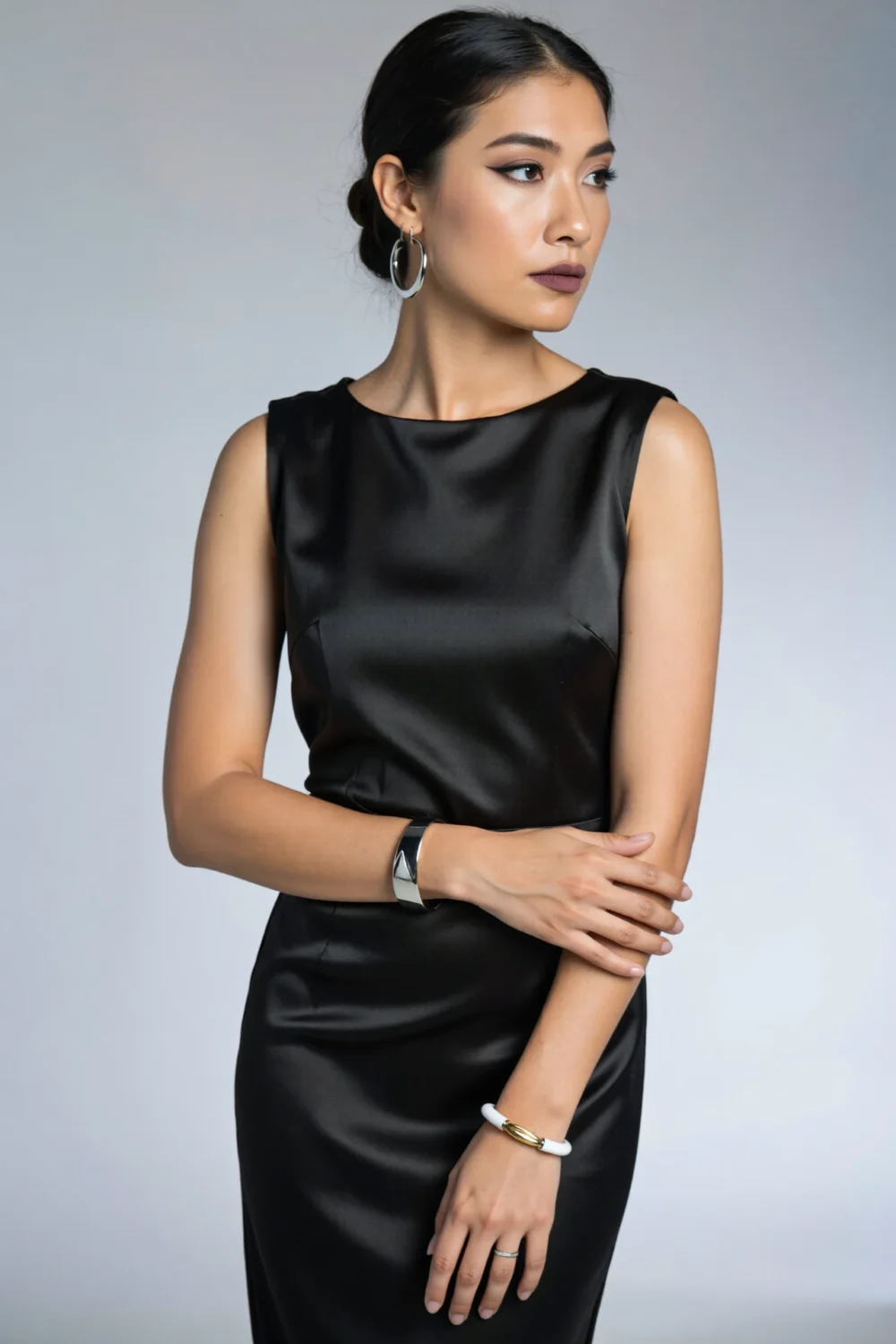 Woman in a satin black LBD styled for work-to-dinner with sculptural silver hoops, a knife-edge bangle and slim metallic bracelet.
