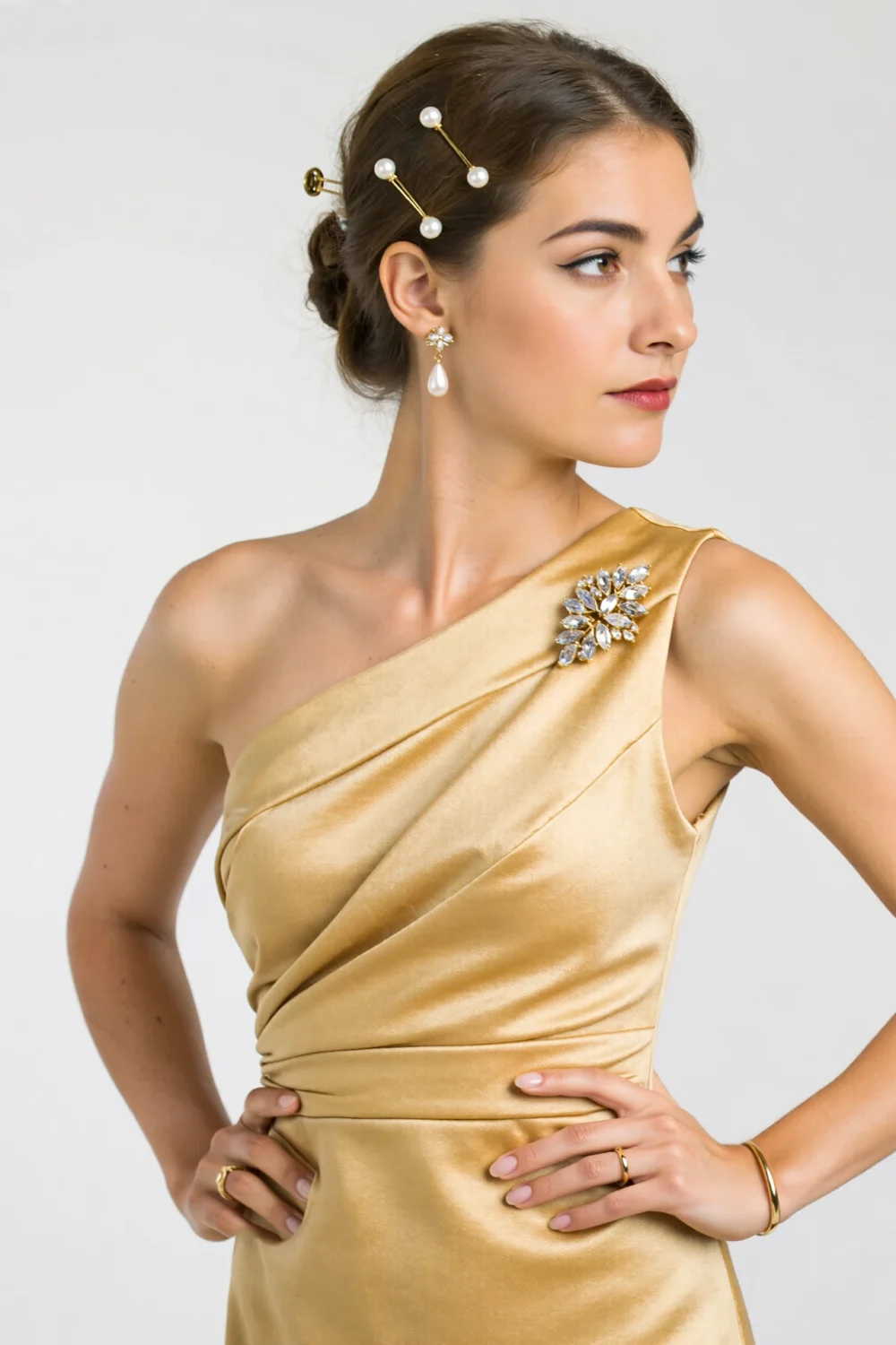 Woman in a gold one-shoulder dress wearing a crystal brooch and pearl hair pins, creating an elegant focal accent