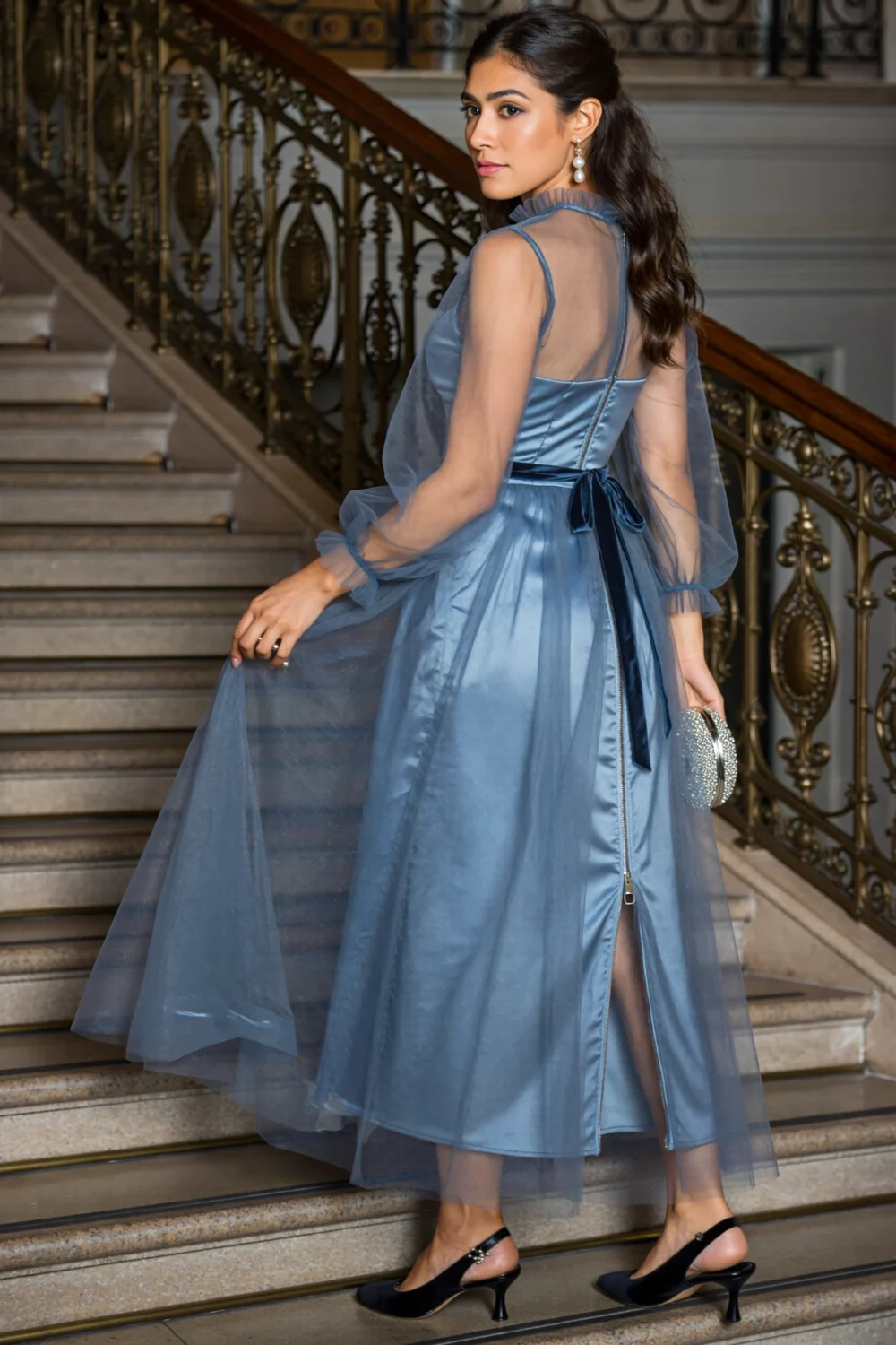 Dusty slate blue tea-length tulle dress with satin bodice, ruffled neckline, puff sleeves, and sheer skirt on staircase; black pumps complete look