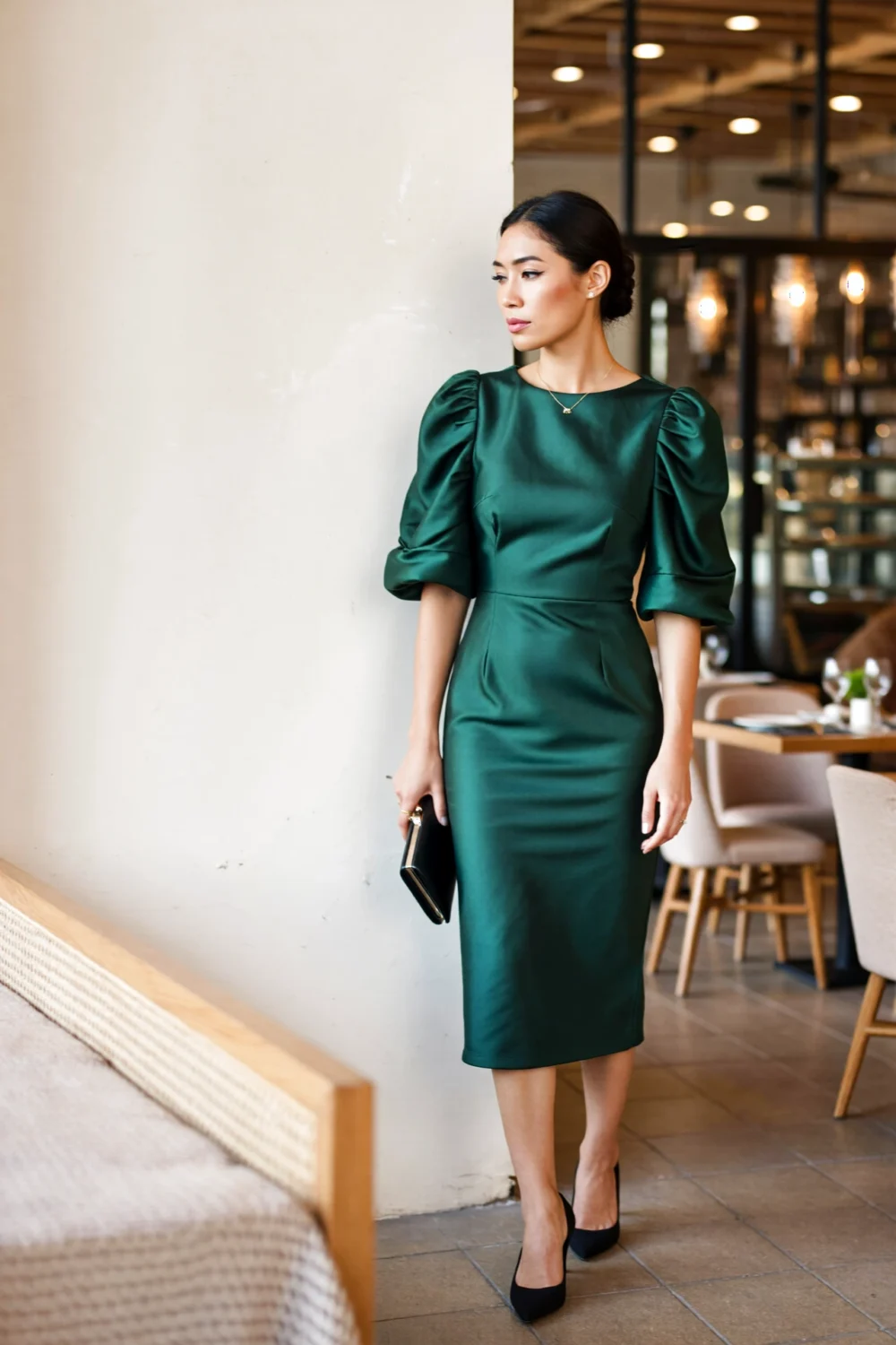Woman in an emerald statement-sleeve midi dress with puff shoulders and a streamlined skirt, clutch in hand, polished dinner-ready