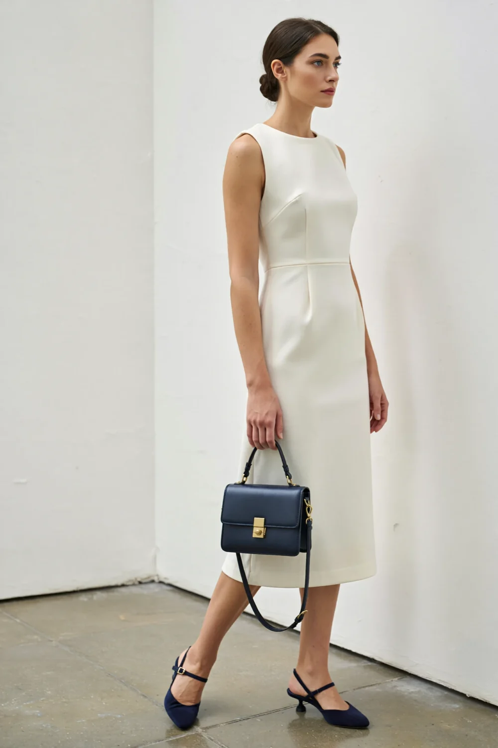 Model in a stone-colored sheath dress holding a structured navy mini bag, paired with navy slingback heels for refined cohesion