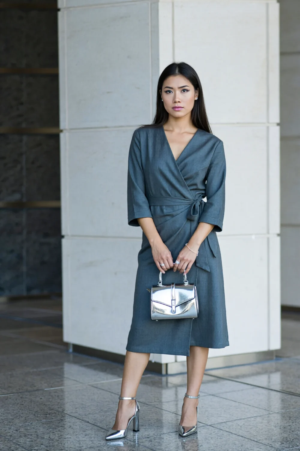 Woman in a stone wrap dress holding a chrome-accented bag, wearing silver block heels and minimal jewelry for sleek polished look
