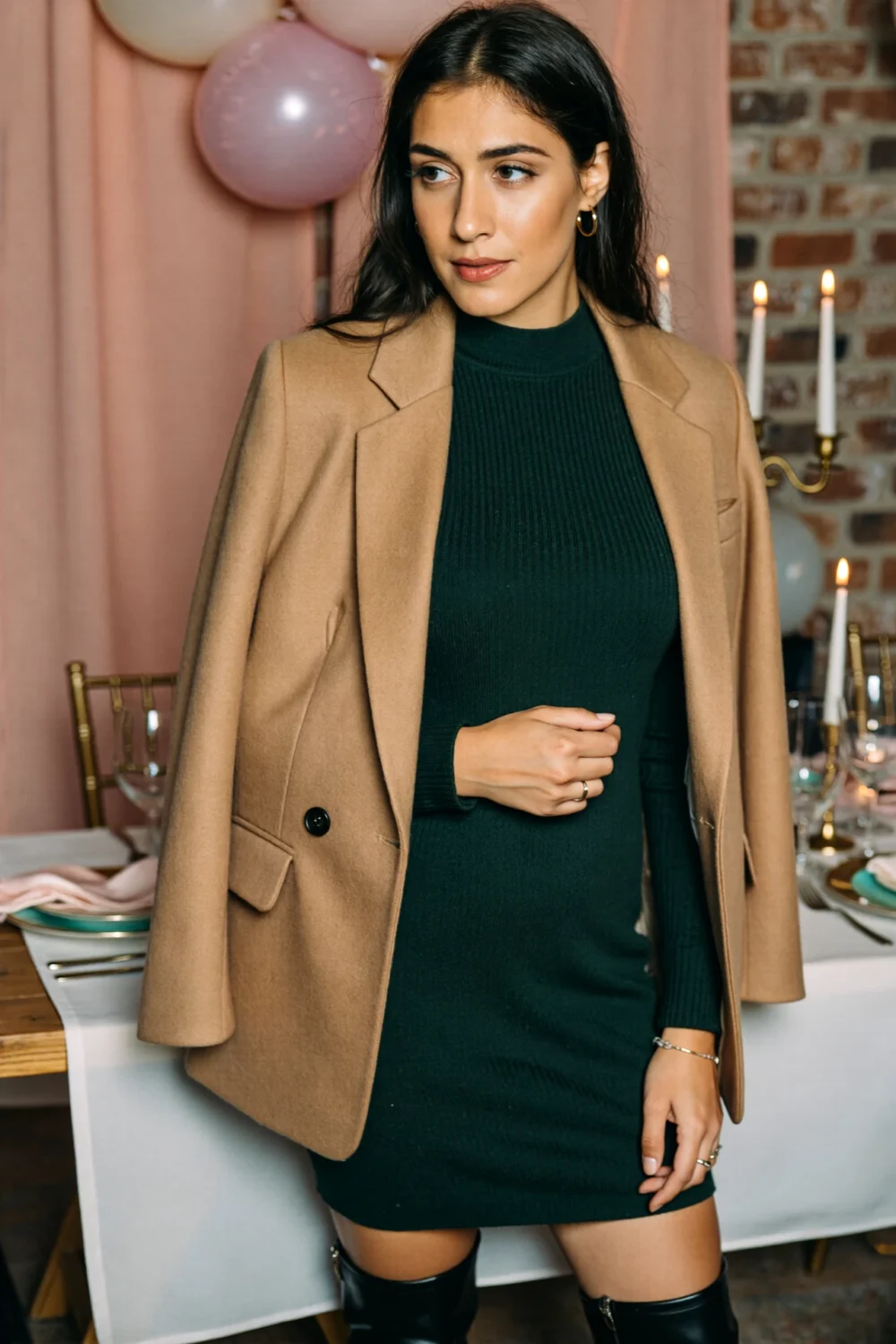 Woman wearing a camel tailored blazer draped over a green knit dress, knee boots and simple jewelry at a candlelit table