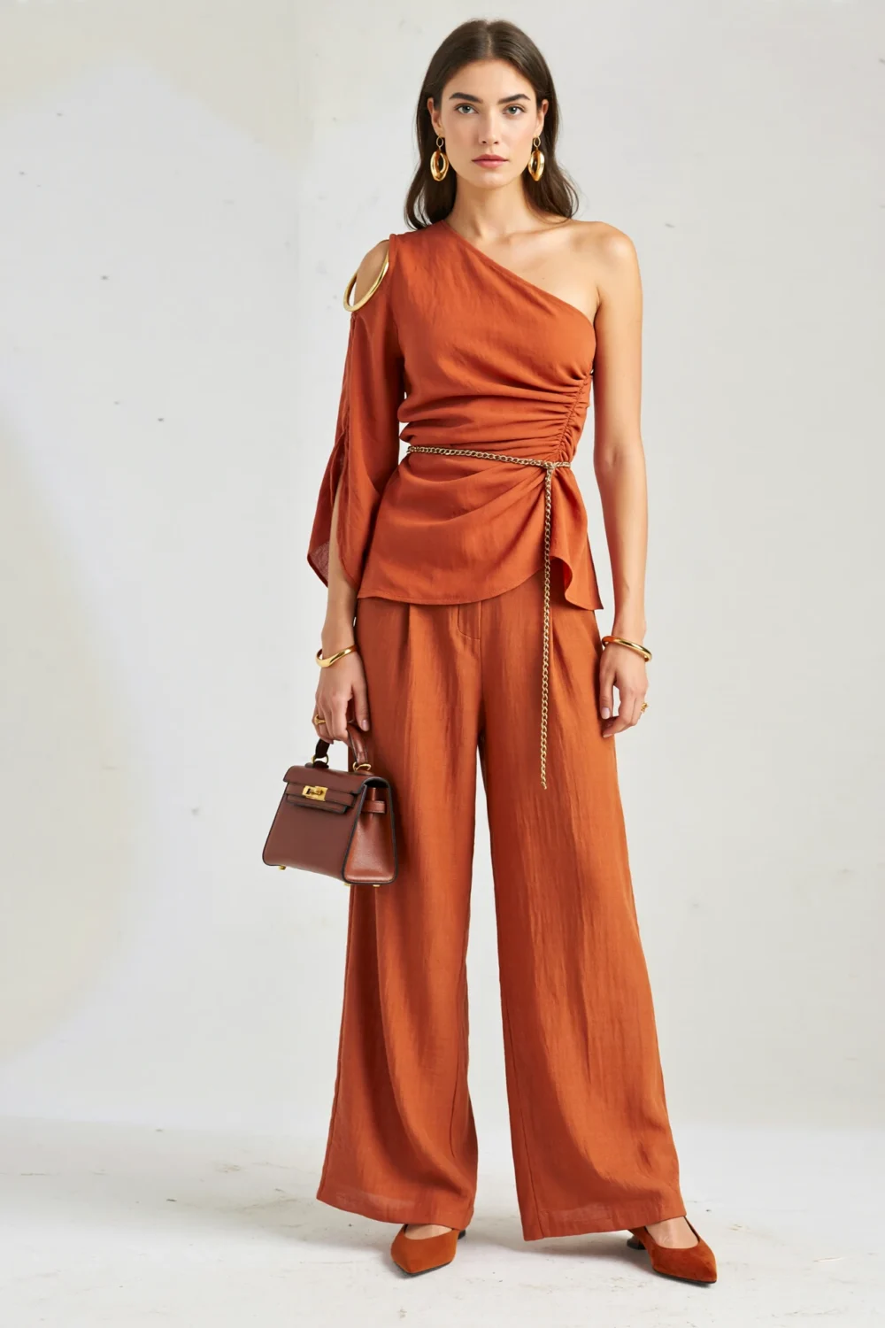 Terracotta asymmetrical one-shoulder top with gold ring, wide-leg pleated trousers, chain belt, and pointed pumps in a chic set