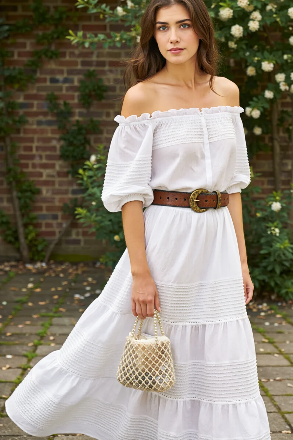 Woman in a tiered white maxi skirt and off-shoulder puff blouse, cinched with a wide belt and holding a beaded bag outdoors
