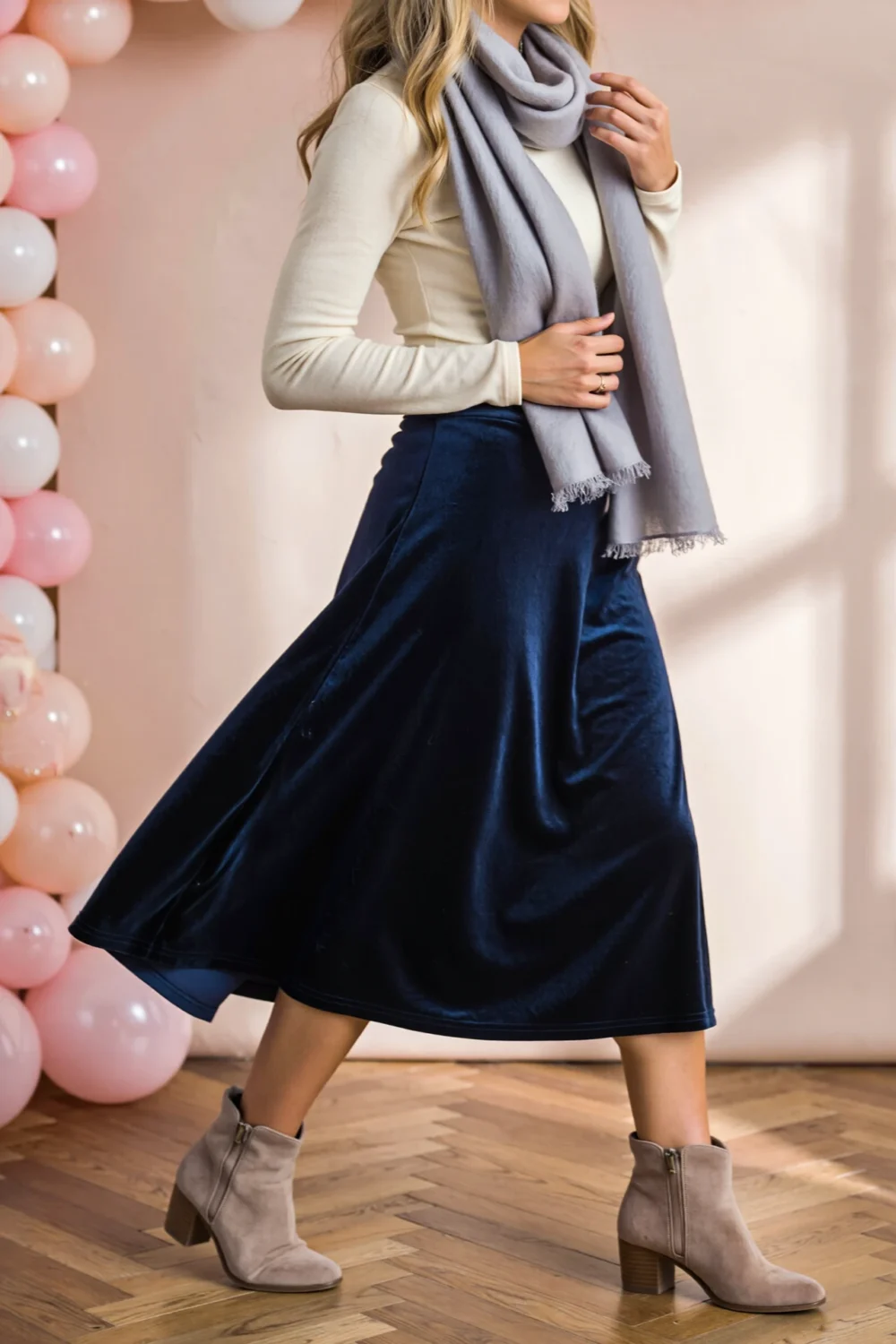 A-line navy velvet midi skirt paired with a light grey linen-texture scarf, cream fitted top and taupe suede booties in motion