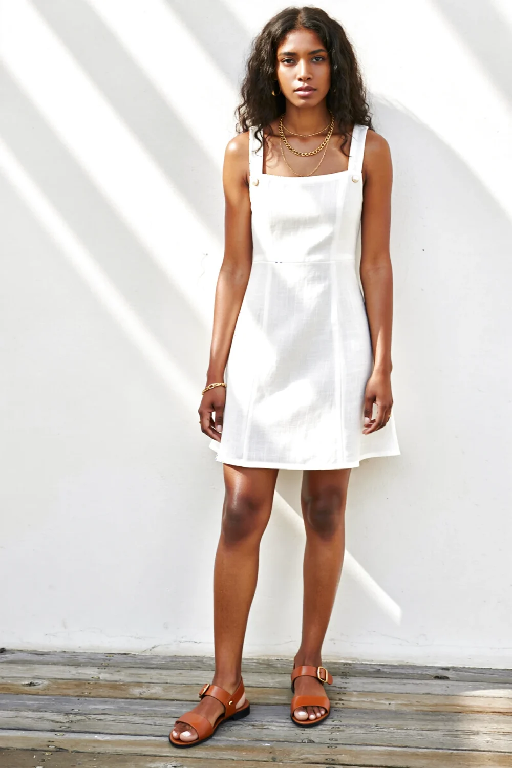 Model in a crisp white mini dress with square neckline, layered gold jewelry and caramel nude-for-you sandals on warm skin in sun.