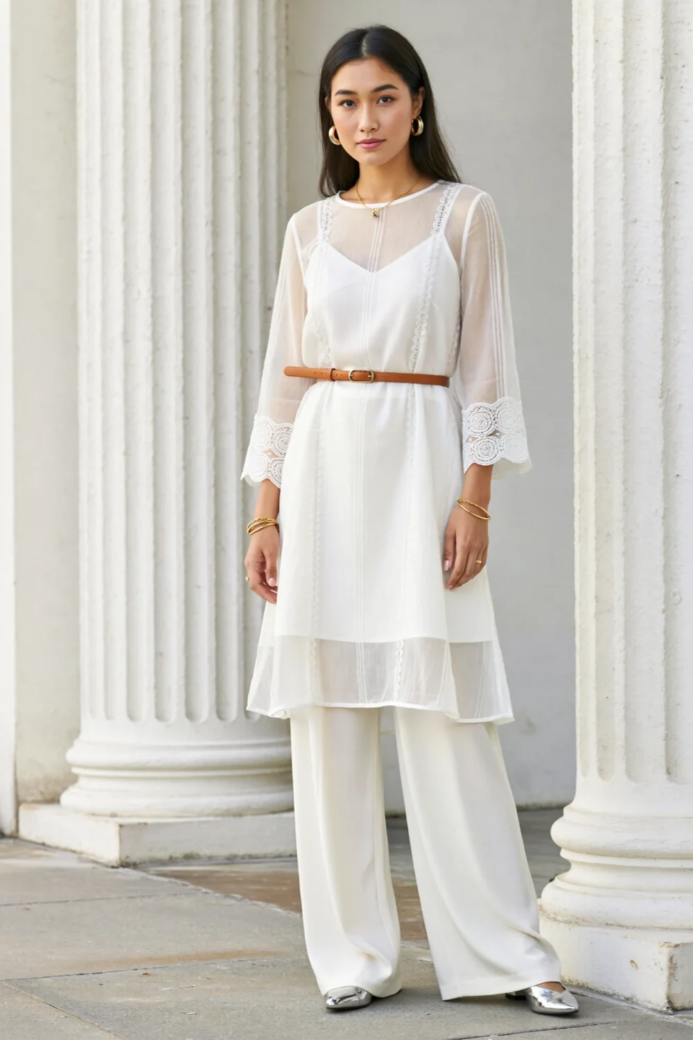 White tunic dress with sheer lace sleeves over wide-leg white trousers, styled with tan belt and metallic shoes in monochrome elegance